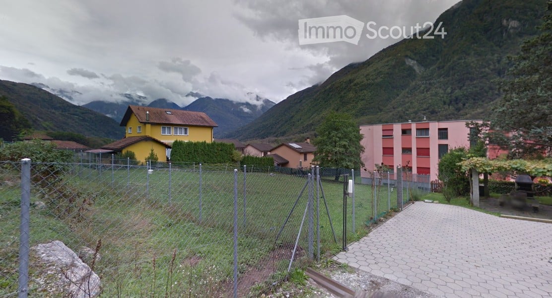A fenced garden area with a paved pathway leading to a residential complex, surrounded by trees and mountains in the background.