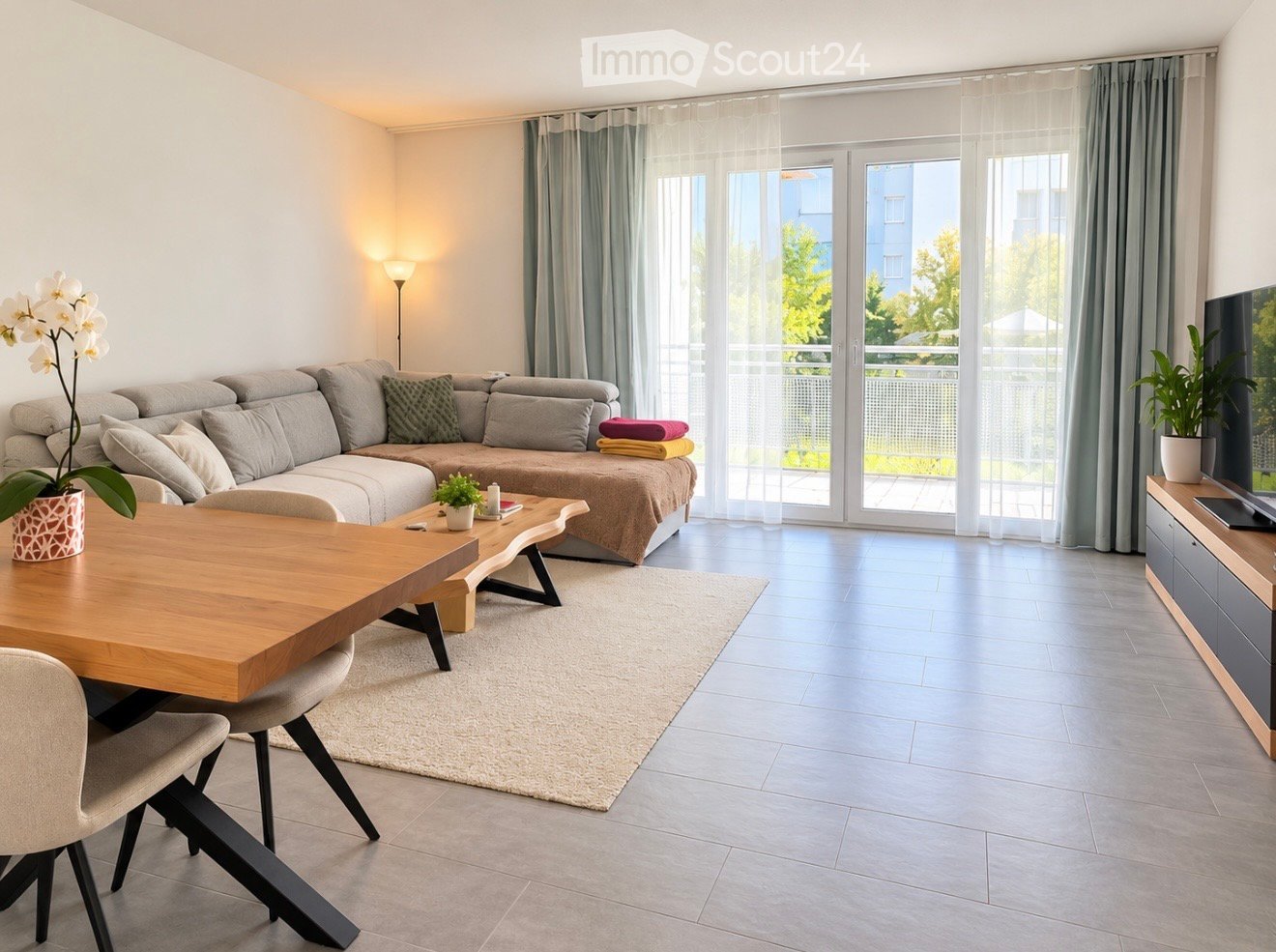 Modern living room with a grey sectional sofa, wooden coffee table, dining table with chairs, potted plants, and a television on a wooden stand. Large windows with sheer curtains lead to a balcony.