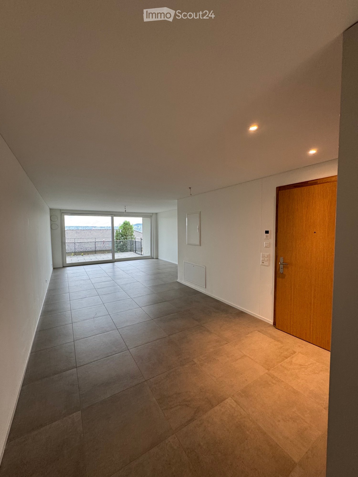 Empty hallway with tiled flooring, white walls, recessed lighting, and a sliding glass door leading to a balcony area.