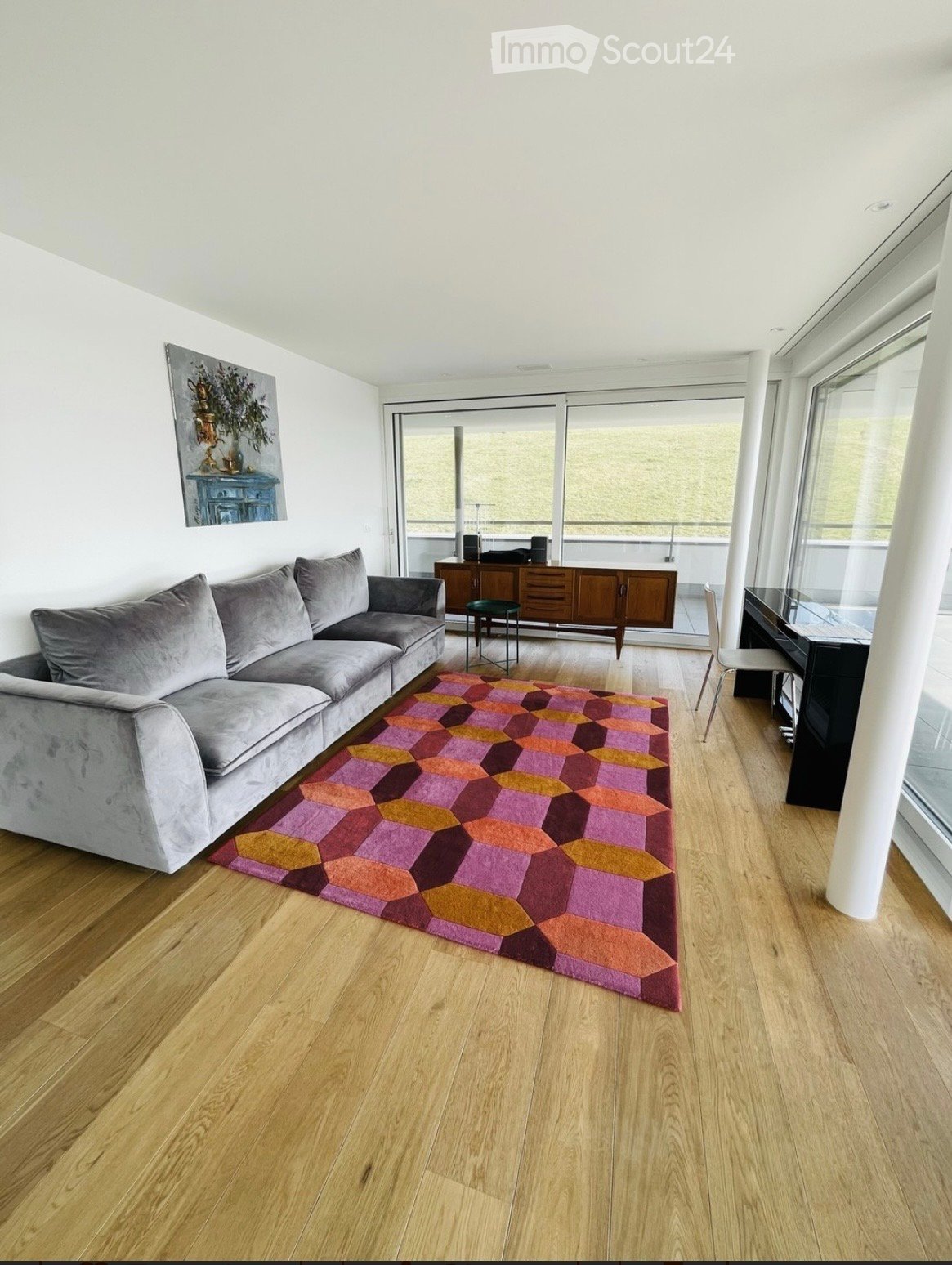 Modern living room with a grey sectional sofa, colorful geometric rug, wooden floor, and large windows leading to a balcony.