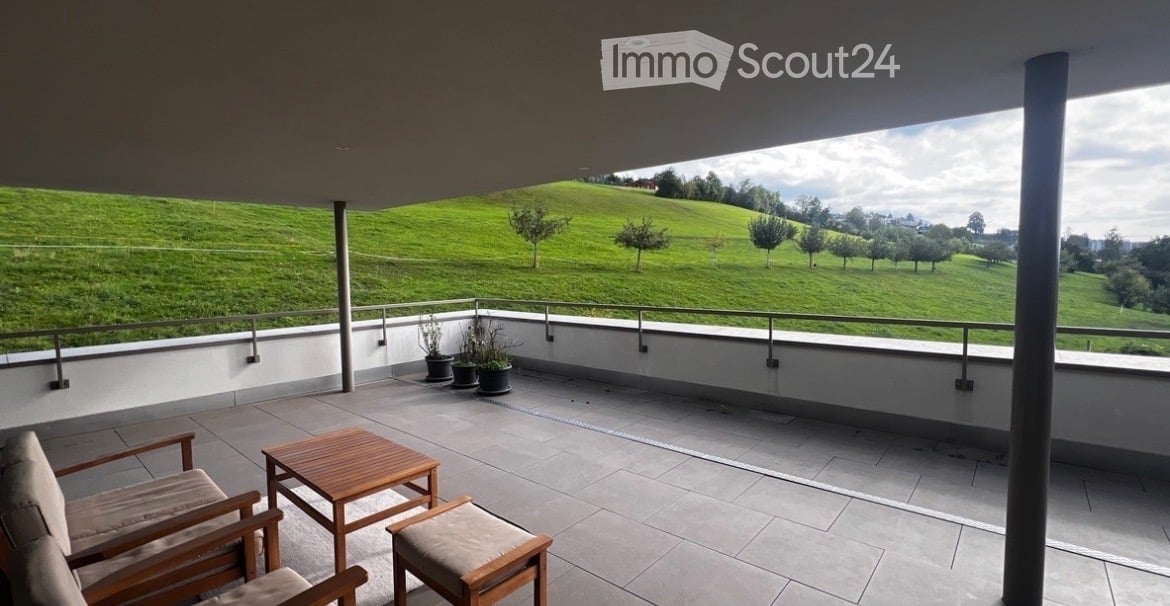 Balcony with a view of green hills, featuring a wooden table, two chairs, a stool, and potted plants.