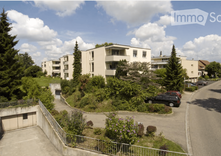 Multi-story residential building with balconies, surrounded by greenery and parking area.