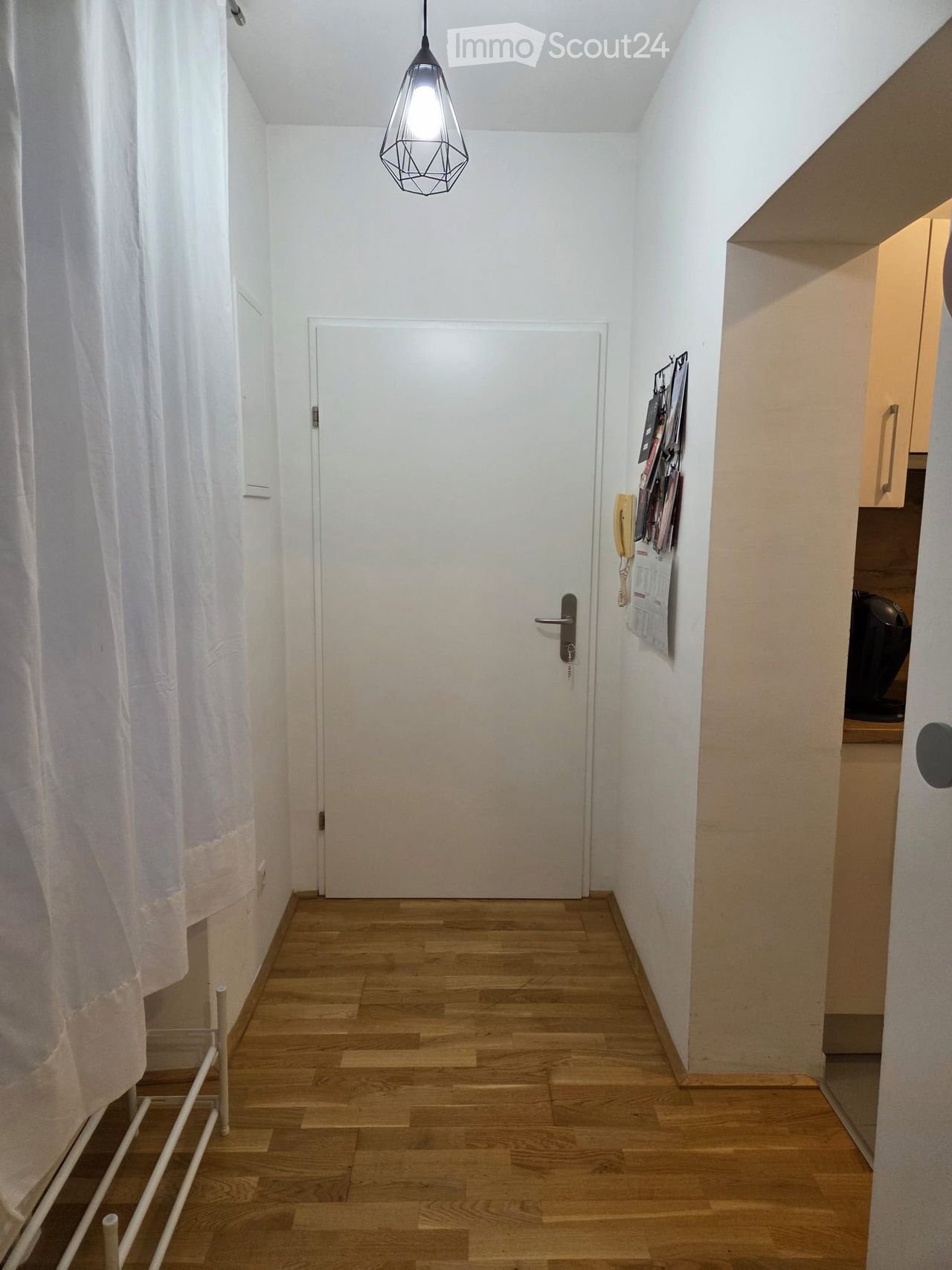 A narrow hallway with wooden flooring, white walls, and a closed door at the end. A hanging light fixture is visible, and a curtain is on the left side.