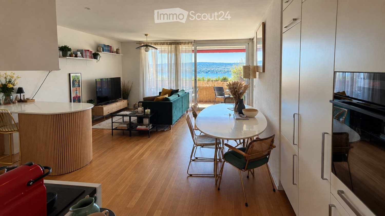 Modern open-plan living and dining area with wooden flooring, large windows offering a scenic view, a sofa, coffee table, dining table with chairs, and a kitchen island.
