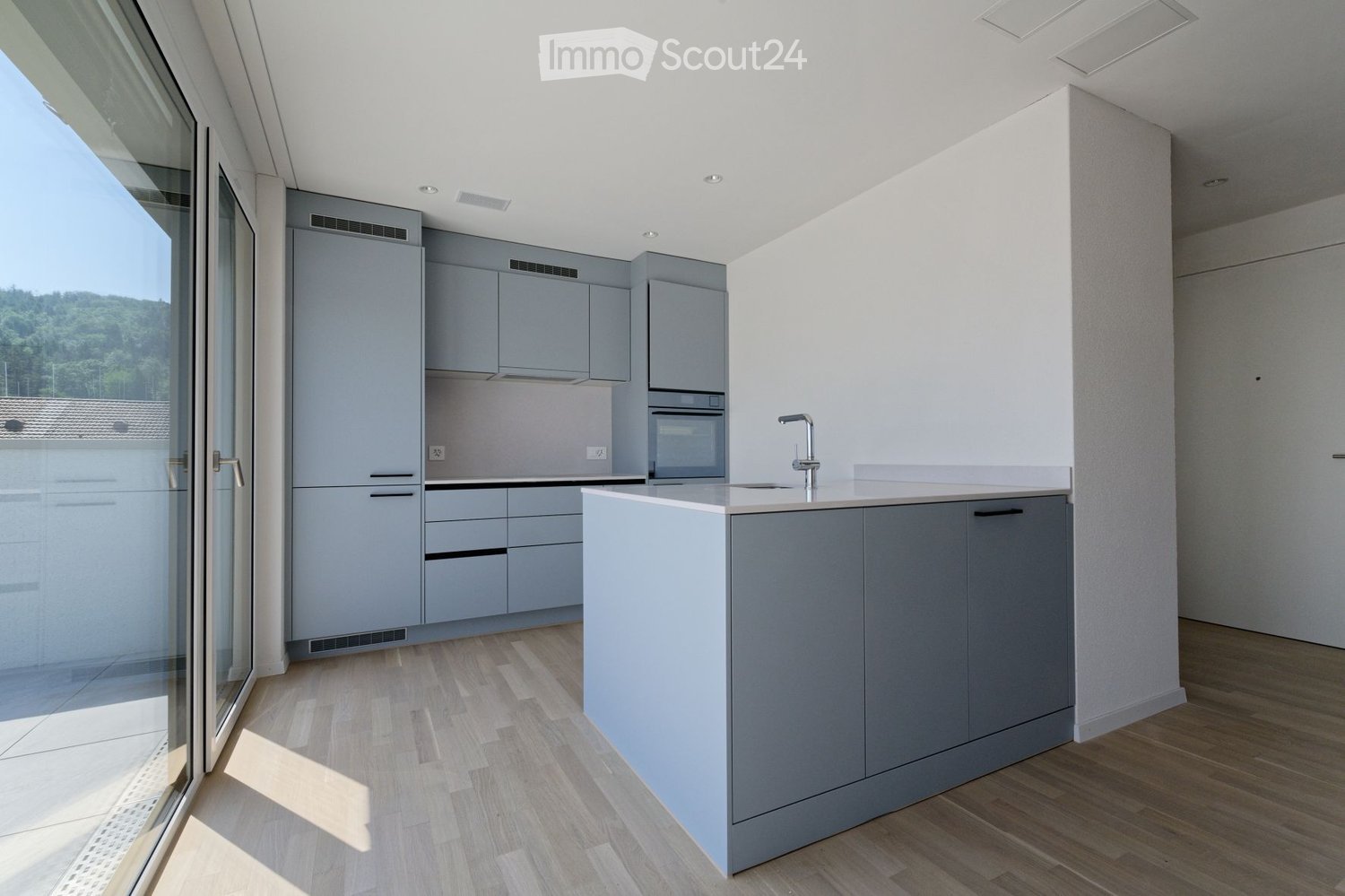 Modern kitchen with grey cabinets, a central island, and a sink with a faucet. The space features wooden flooring and large windows offering a view of the outdoors.