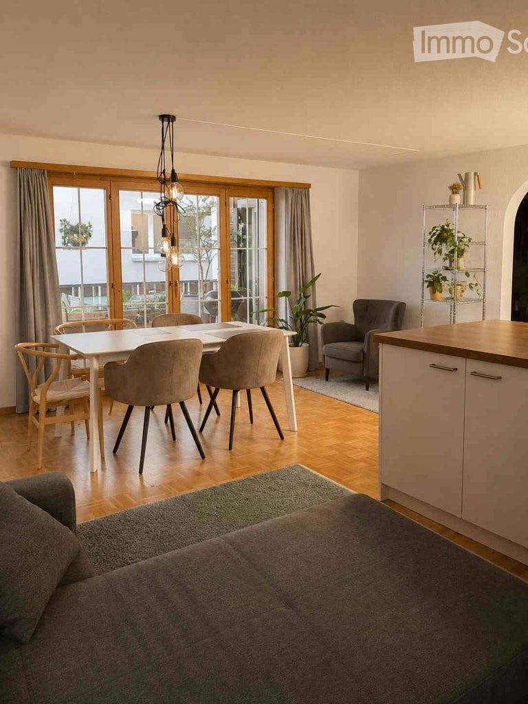 Modern open-plan living and dining area with a grey sofa, dining table with chairs, kitchen cabinetry, potted plants, and a shelf with decorative items.