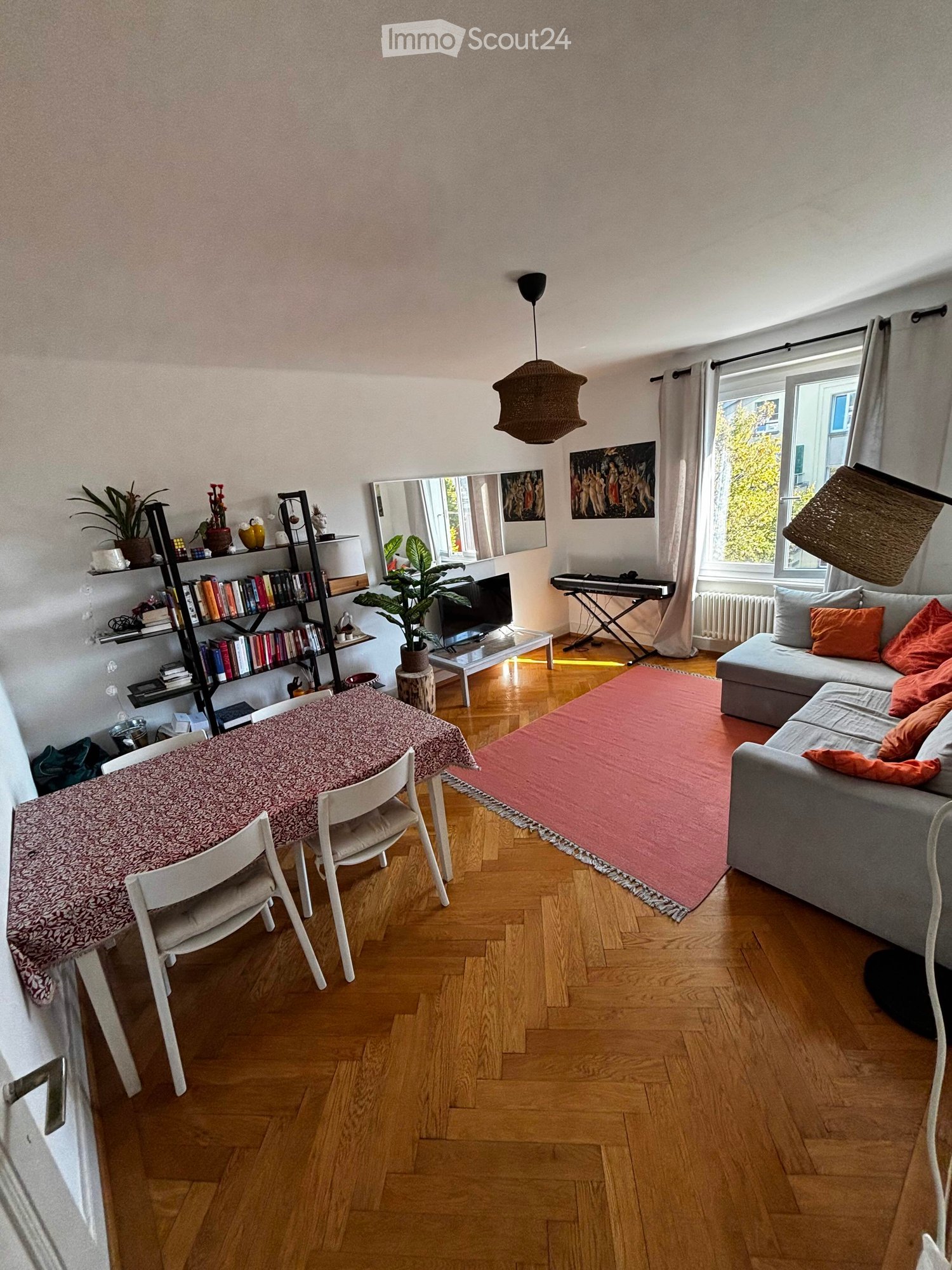 A modern living room with wooden flooring, a grey sofa with orange cushions, a pink rug, a dining table with chairs, a bookshelf, a piano, and a large mirror. Natural light streams in from large windows.