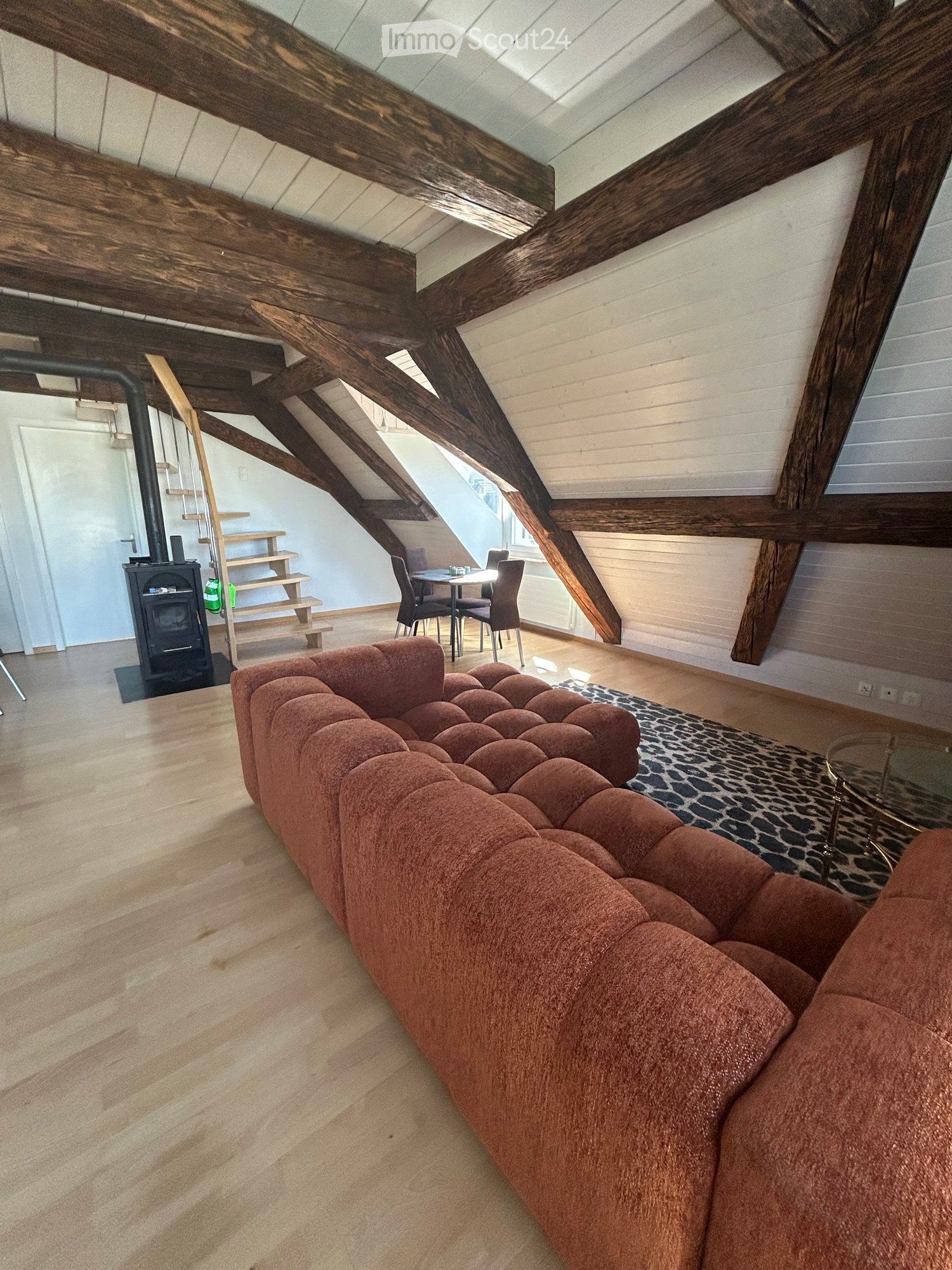 Attic living space with exposed wooden beams, a red tufted sofa, a glass coffee table, a dining area with chairs, and a fireplace.