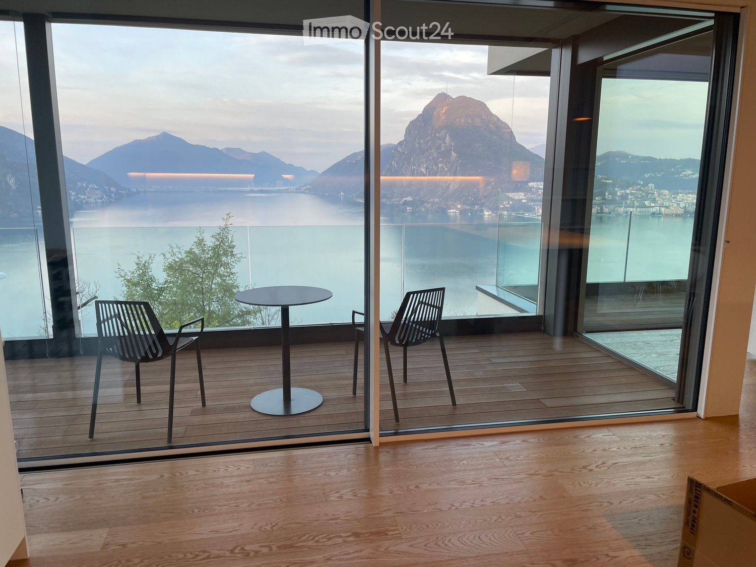 Modern balcony with wooden flooring, large glass walls offering a scenic view of a lake and mountains, equipped with a round table and two contemporary chairs.