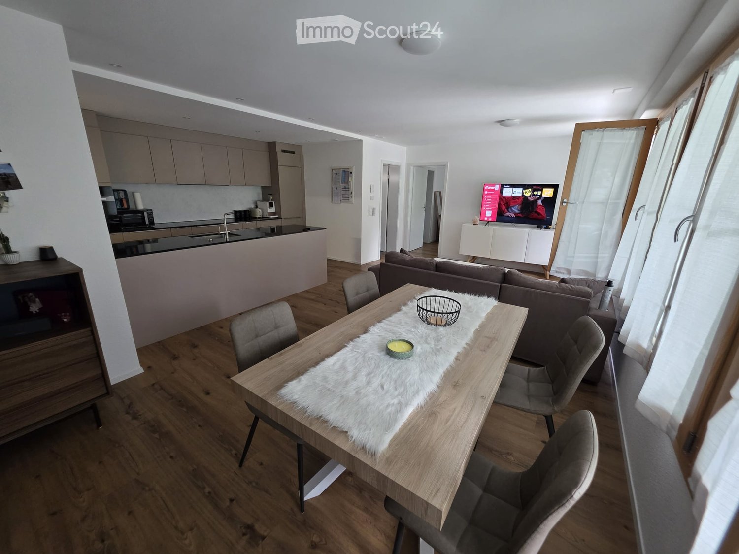 Open-plan living and dining area with a sofa, dining table, chairs, and a television. The kitchen area features a microwave and cabinets.
