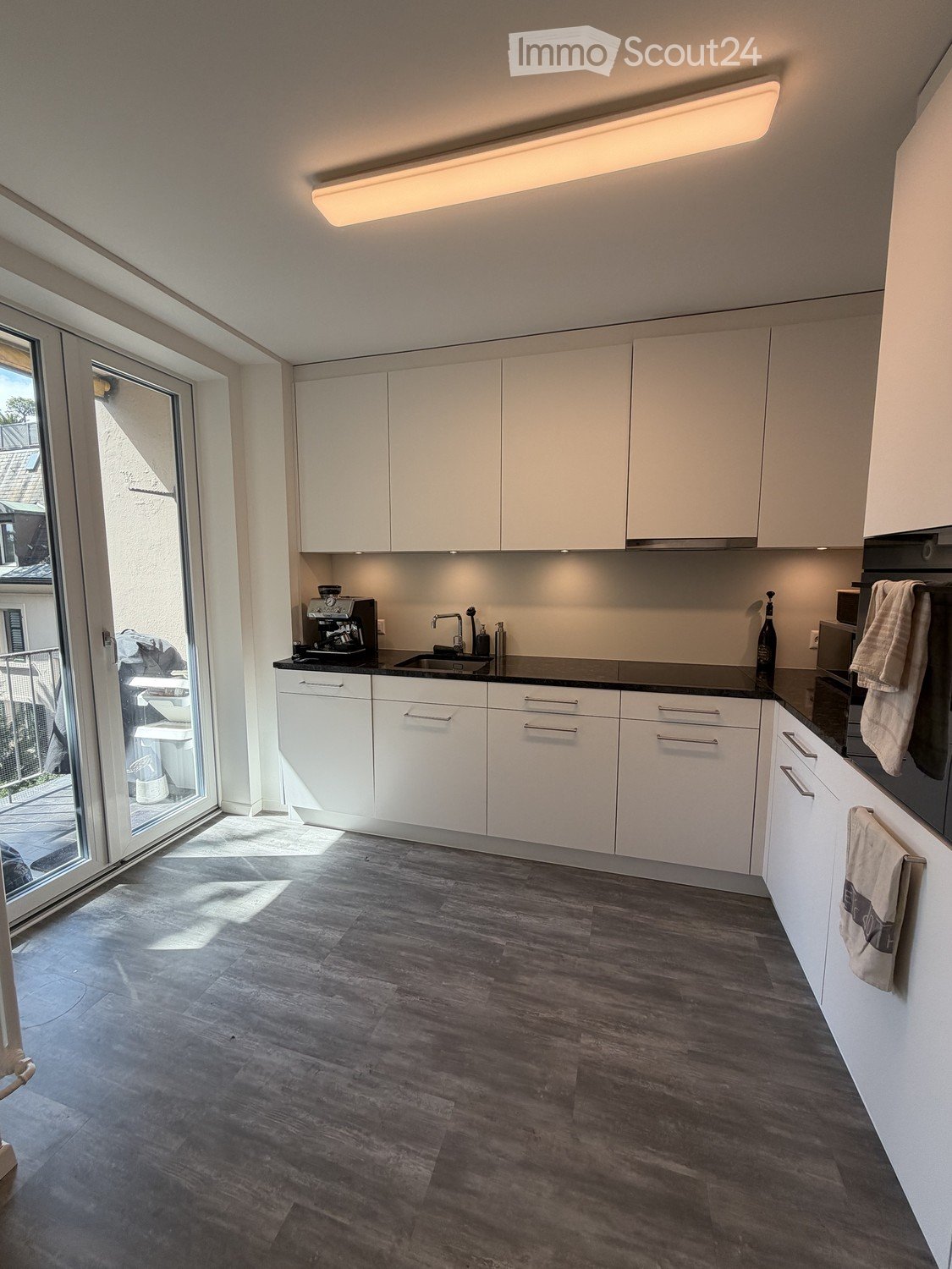 Modern kitchen with white cabinets, dark countertops, and wooden flooring. Features a coffee maker, sink, and oven.