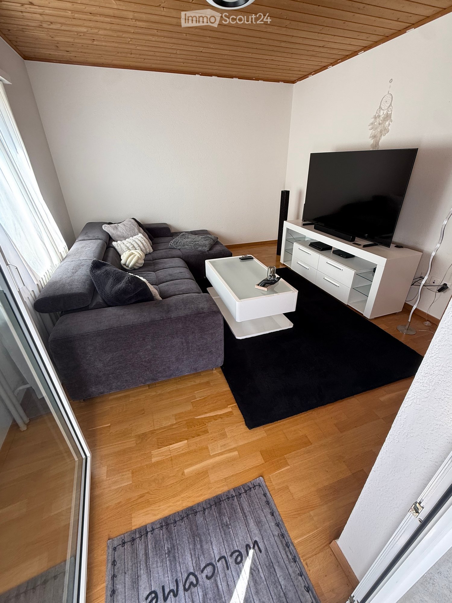 Modern living room with a grey sectional sofa, white coffee table, TV stand with a large flat-screen TV, and wooden flooring.