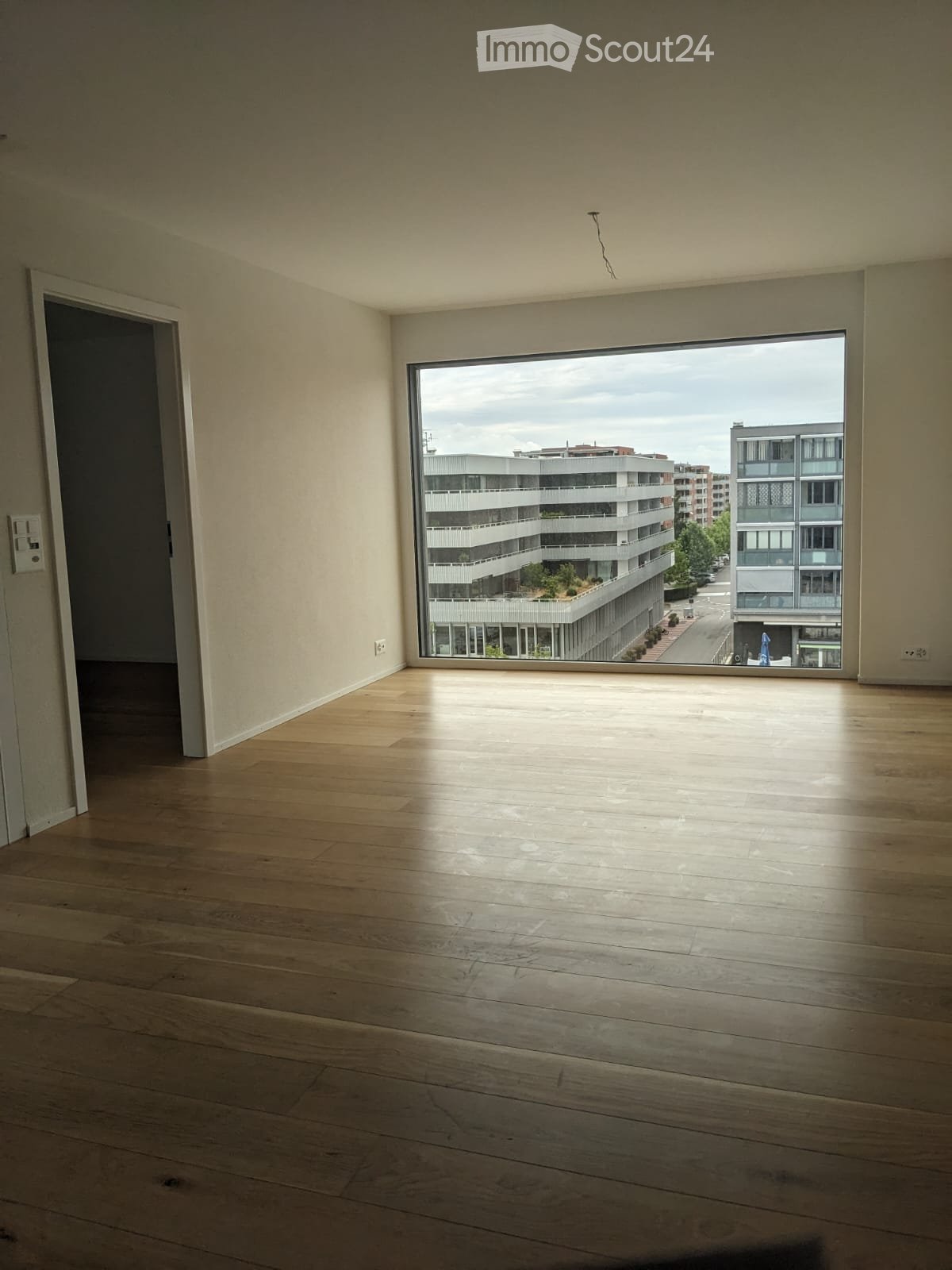 Empty room with wooden flooring, large window showing cityscape, and a door leading to another space.