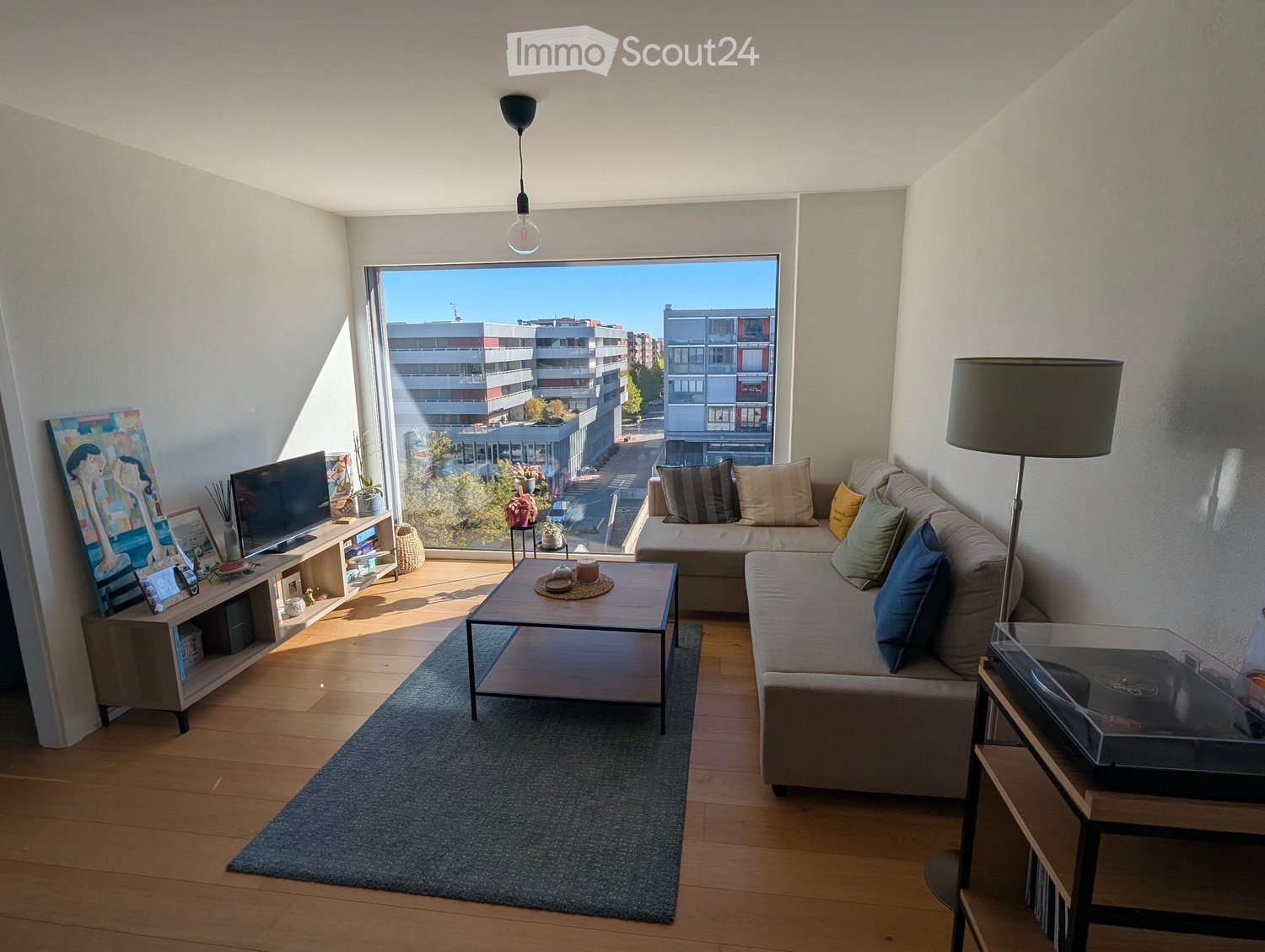 Modern living room with large windows offering a city view, featuring a grey sofa with multiple cushions, a wooden coffee table, a floor lamp, a TV stand with a flat-screen TV, and a painting on the wall.