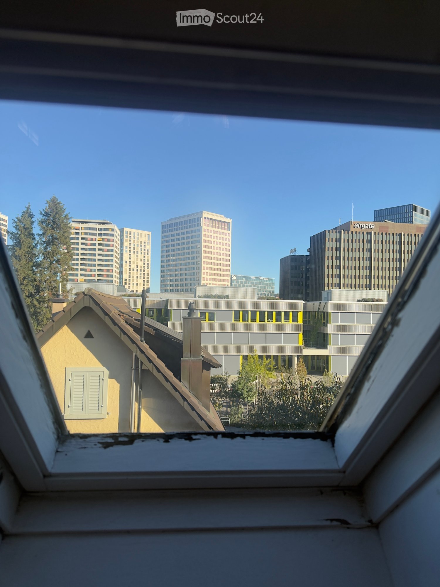 View from a window showing a mix of residential and commercial buildings, including a yellow house with a chimney and several tall office towers in the background.