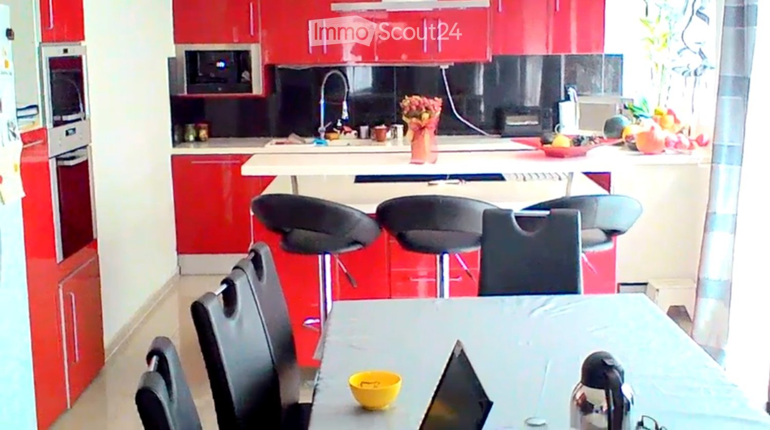 Modern kitchen with red cabinets, white countertops, and stainless steel appliances. Includes a dining area with a table, chairs, and stools.