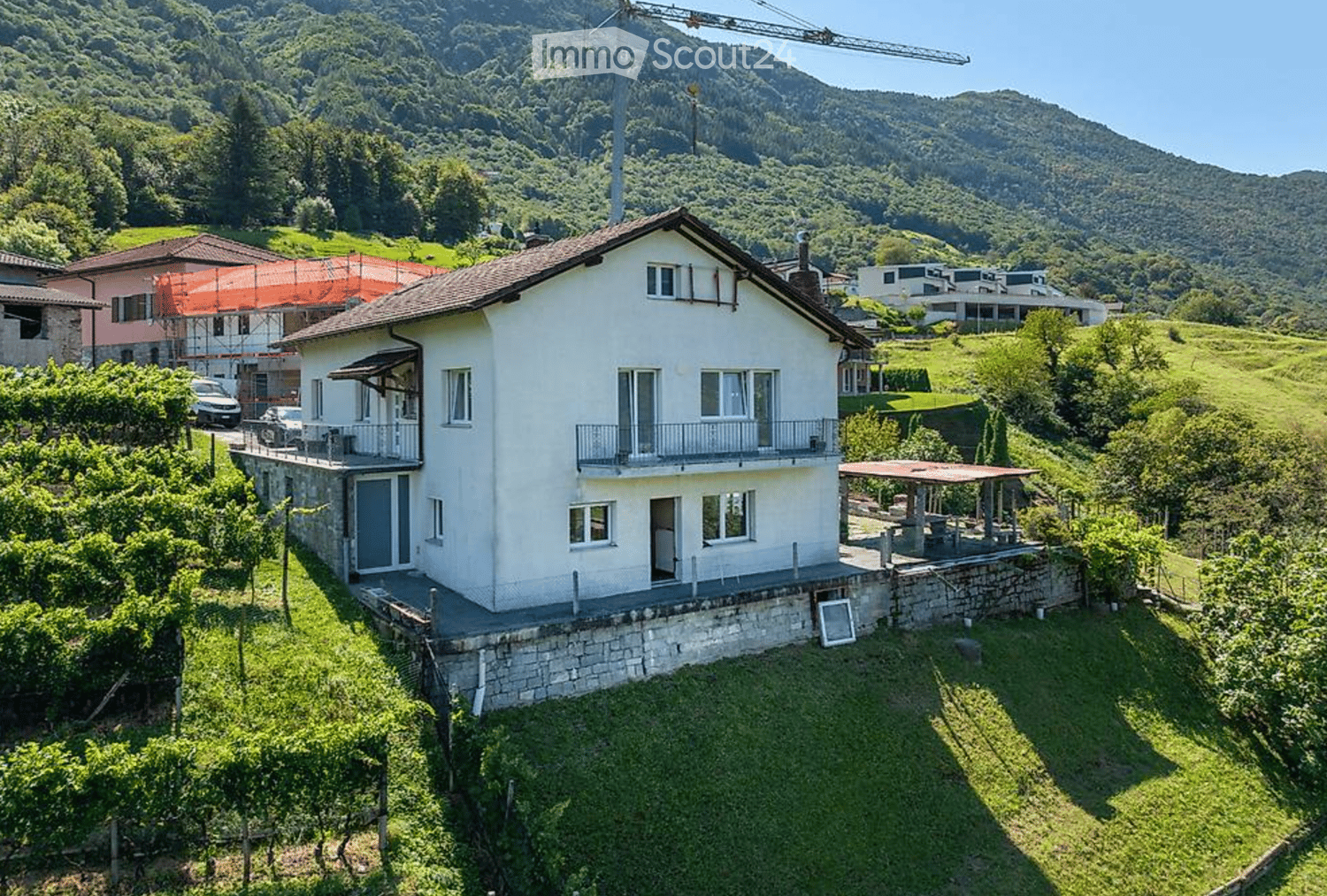 Two-story white house with balconies, surrounded by greenery and vineyards, with a scenic mountain backdrop.