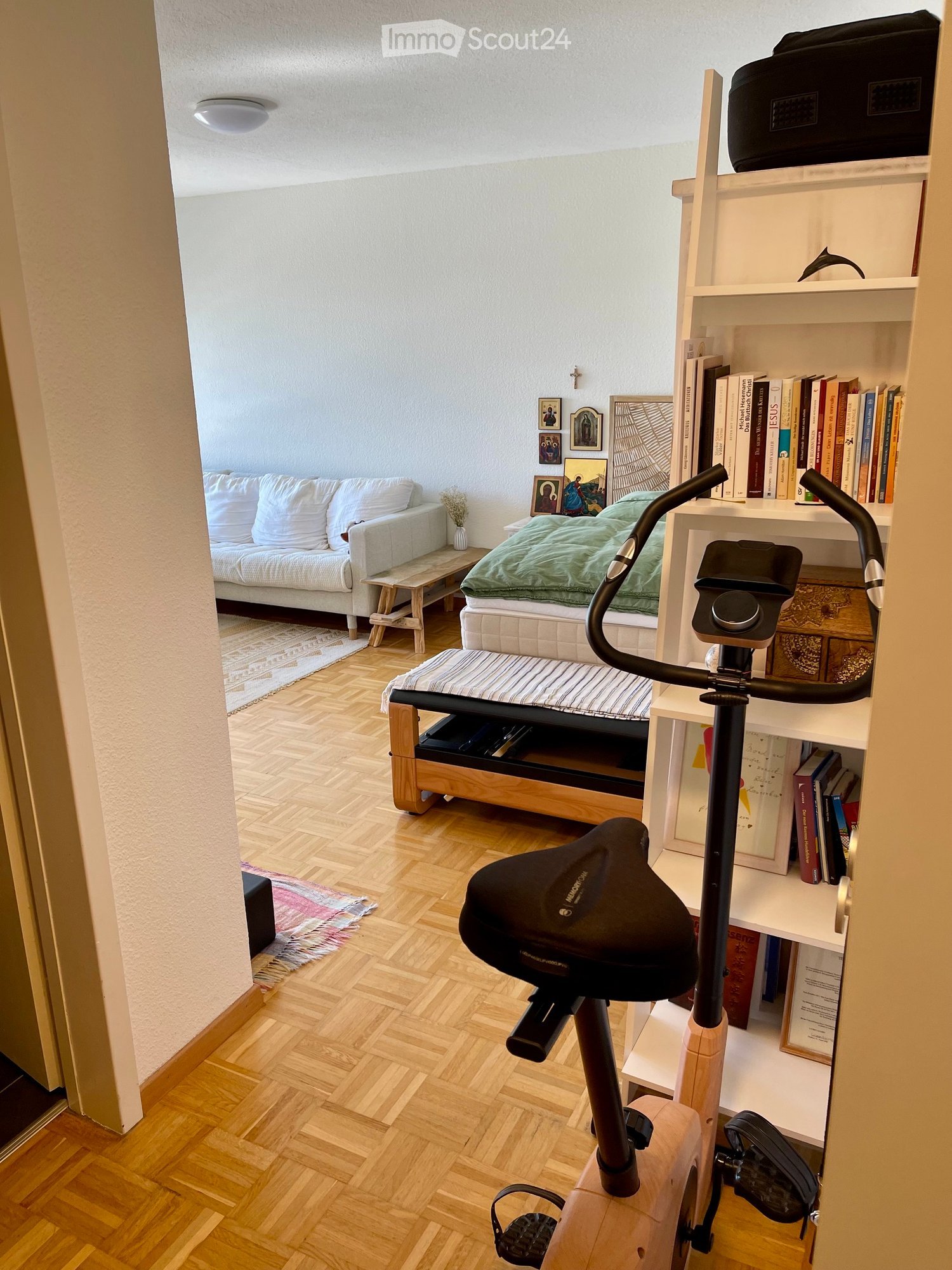 A multi-functional room with a sofa, small table, bed, bookshelf, and exercise bike. The room features wooden flooring and white walls.