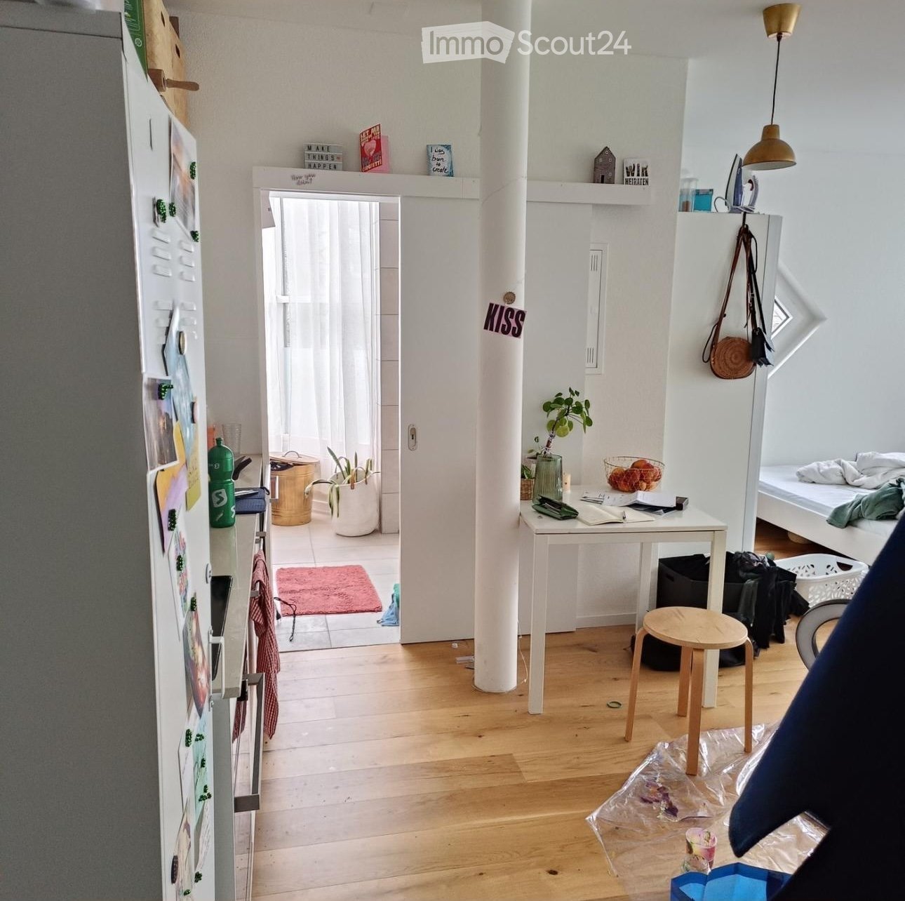 A modern, minimalist living space with light wooden flooring, white walls, and a mix of functional and decorative items. Visible objects include a small wooden stool, a white table with a bowl of fruit, a refrigerator with magnets and notes, a potted plan
