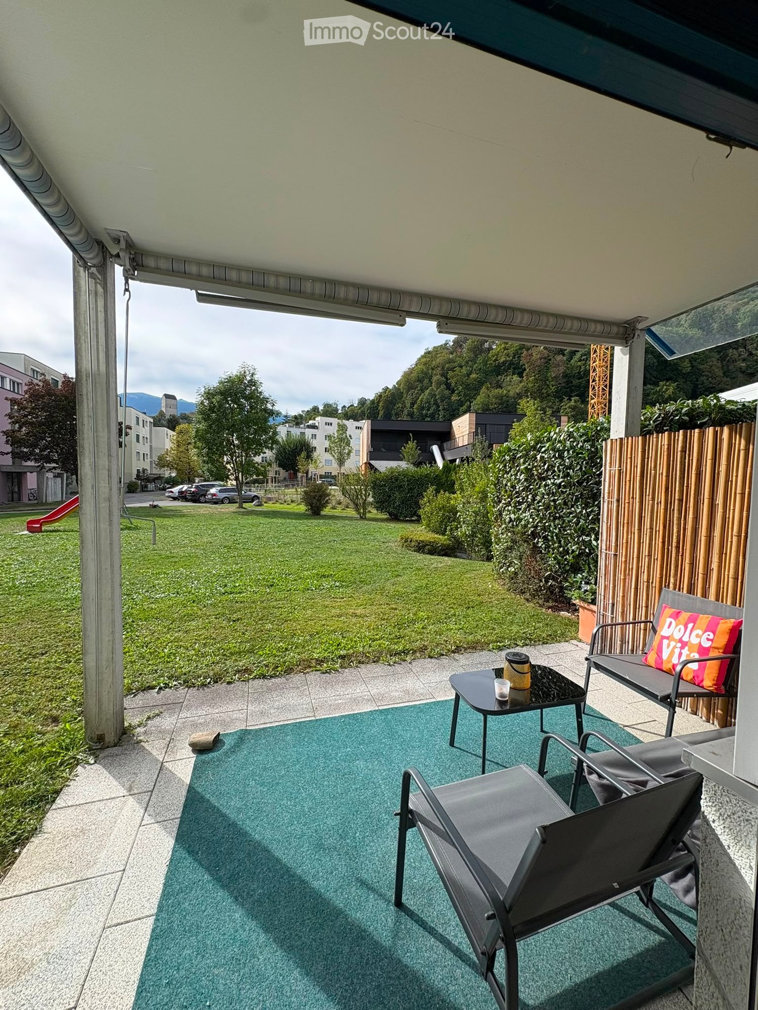 Balcony with a green rug, two chairs, a small table, and a cup on the table. The view shows a grassy area with trees and buildings in the distance.