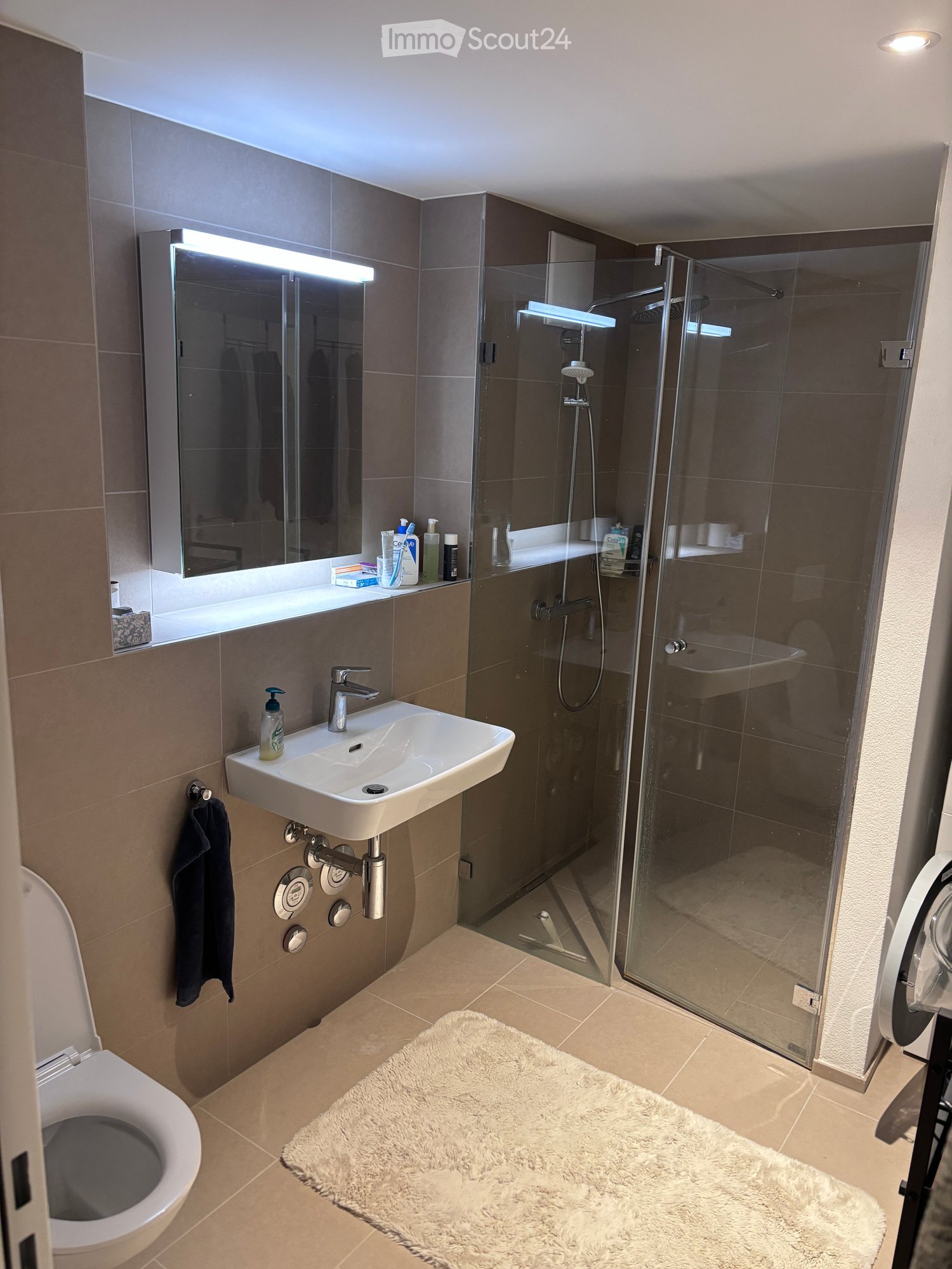 Modern bathroom with a white sink, toilet, large mirror, glass-enclosed shower, and a fluffy white rug on the floor.
