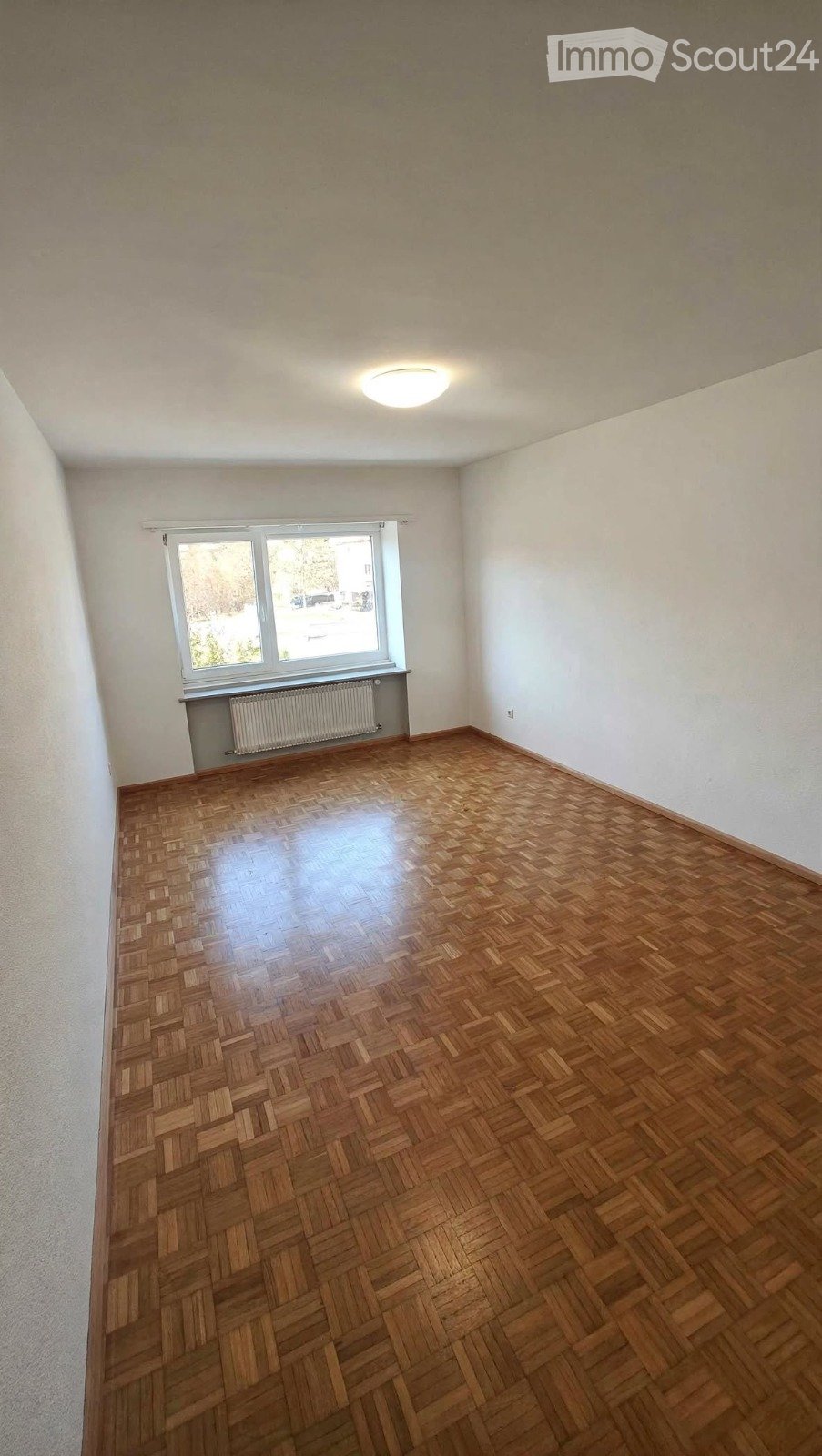 Empty room with wooden parquet flooring, white walls, a large window with a radiator below, and a ceiling light fixture.