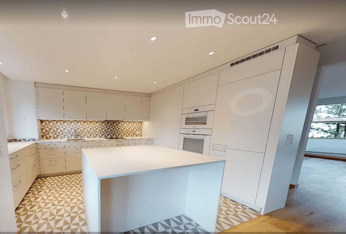 Modern kitchen with white cabinets, a central island, and built-in ovens. The backsplash features a geometric pattern, and the floor has a mix of wood and tile.