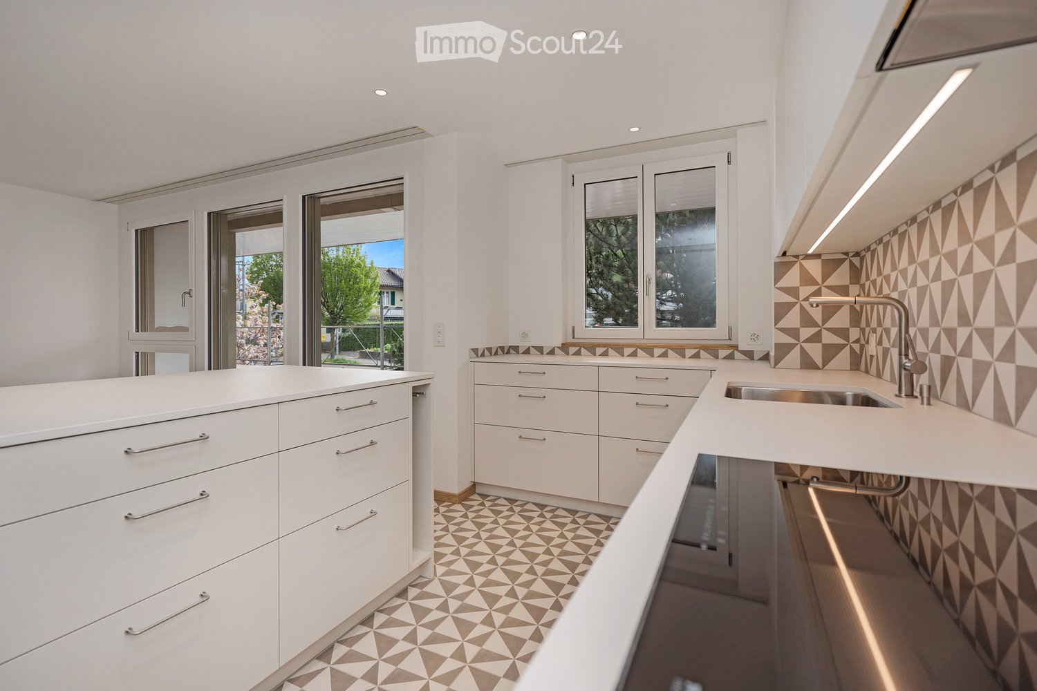 Modern kitchen with white cabinets, stainless steel sink, geometric patterned tiles, and large windows offering a view of greenery.