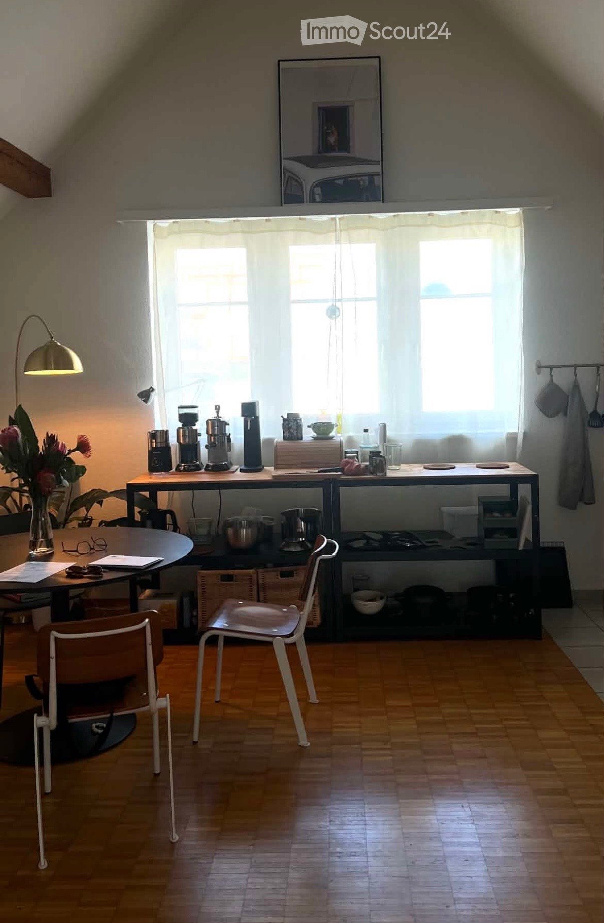 Bright kitchen with wooden flooring, large windows, coffee maker, table, chairs, and decorative vase with flowers.