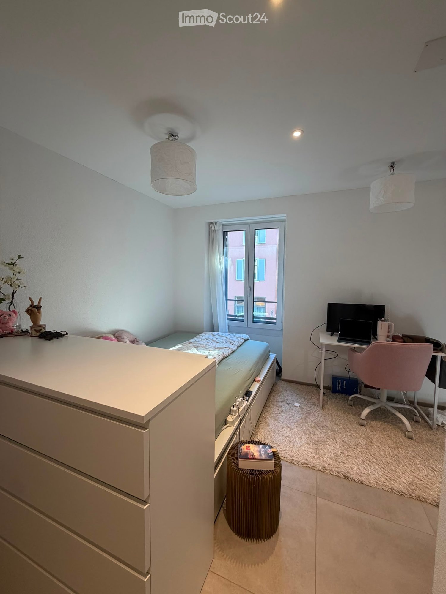A well-lit bedroom with a bed, desk, chair, laptop, and a radiator. The room features a window with a view, a small shelf with decorative items, and a cozy carpeted area.