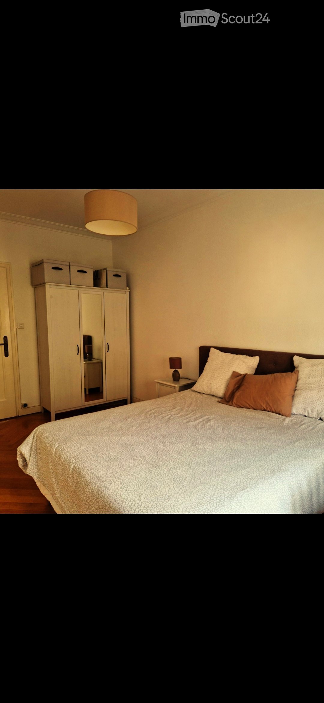 A well-lit bedroom with a large bed, nightstand, lamp, wardrobe, and mirror. The room features wooden flooring and neutral-colored walls.