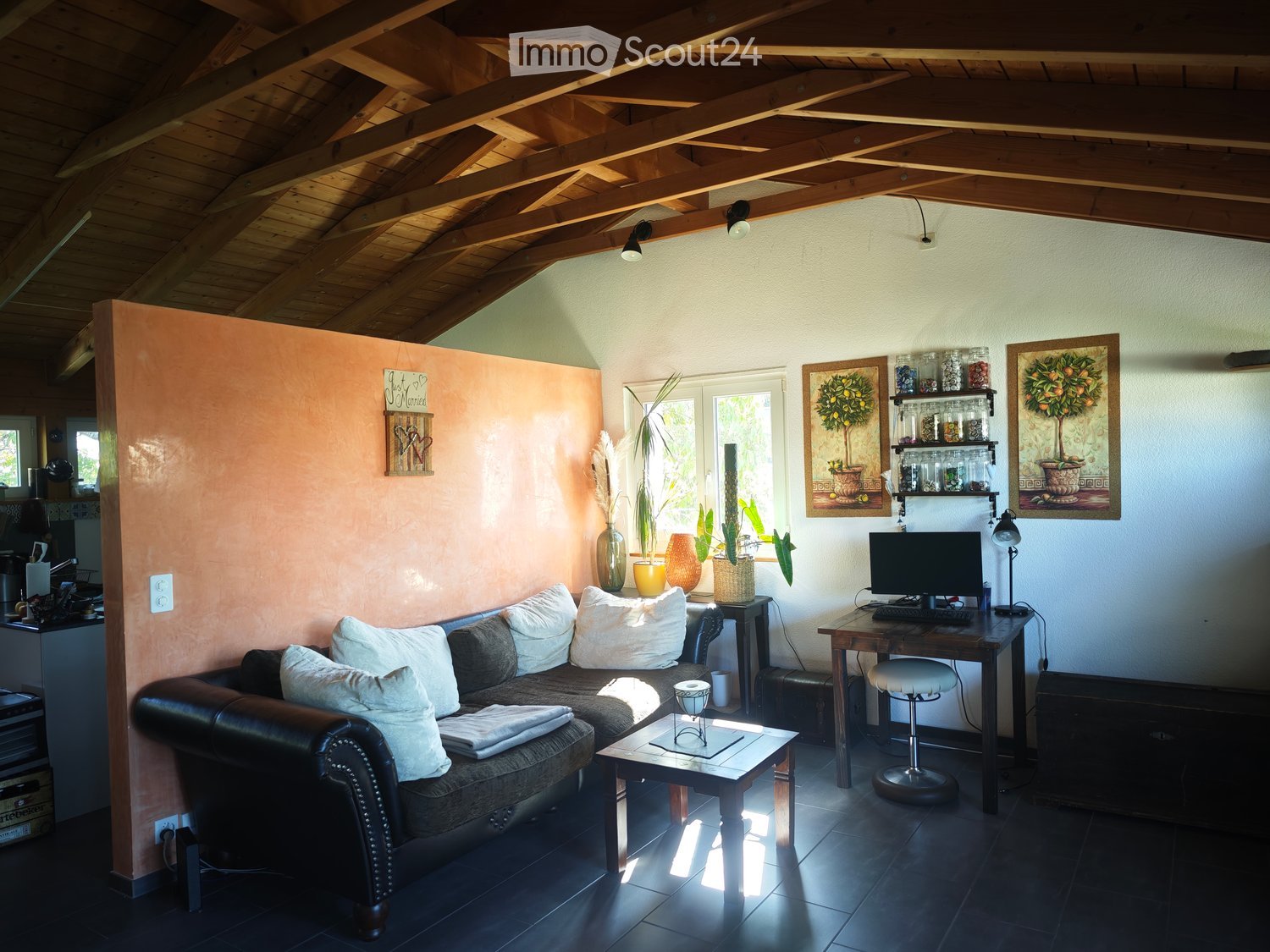 Cozy living room with exposed wooden ceiling beams, dark tiled floor, leather sofa with white cushions, coffee table, desk with computer, and decorative vases.