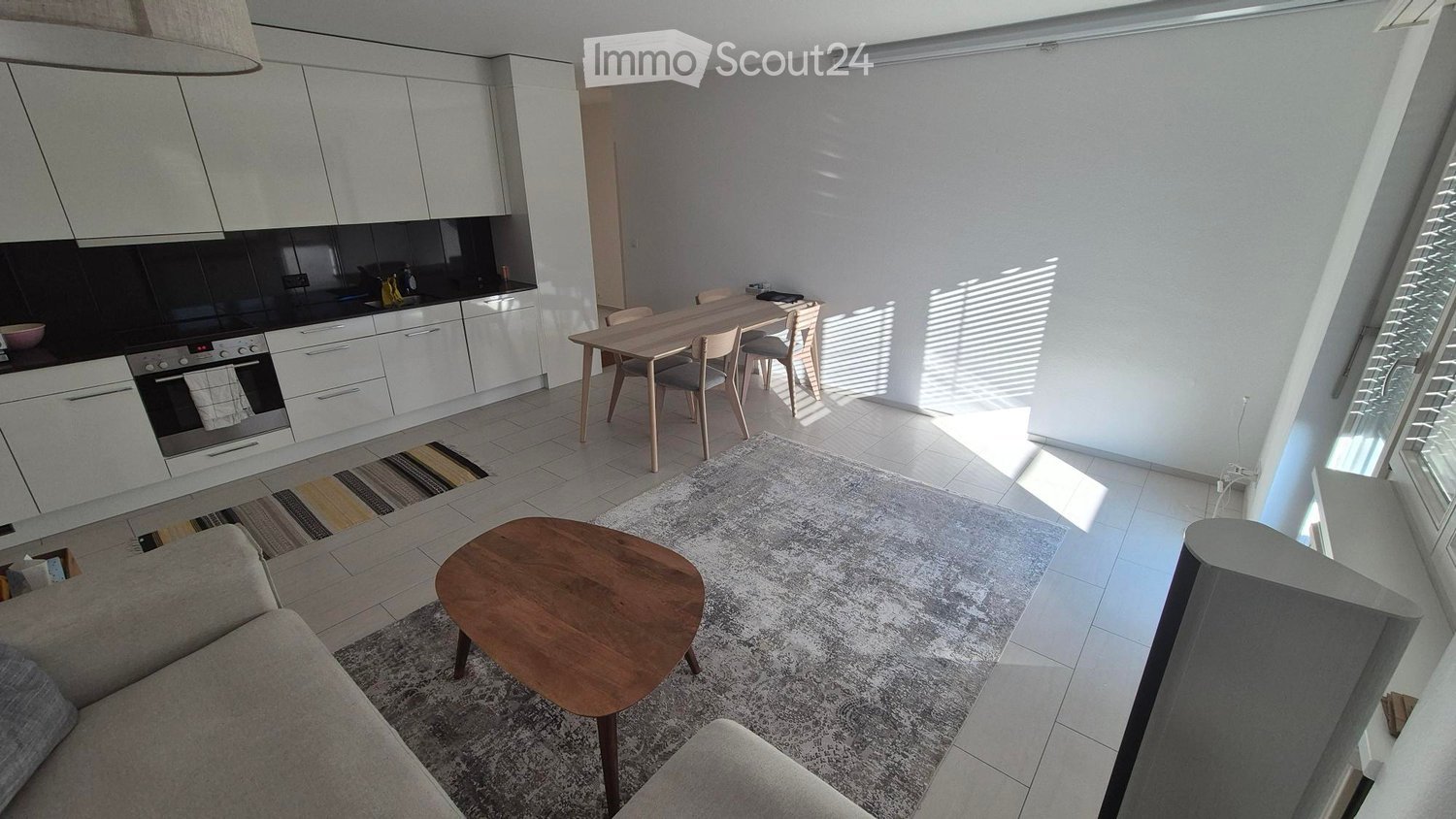 Open-plan living and dining area with modern white kitchen cabinets, a grey sofa, a wooden coffee table, and a dining table with chairs. The space features large windows allowing natural light to fill the room.