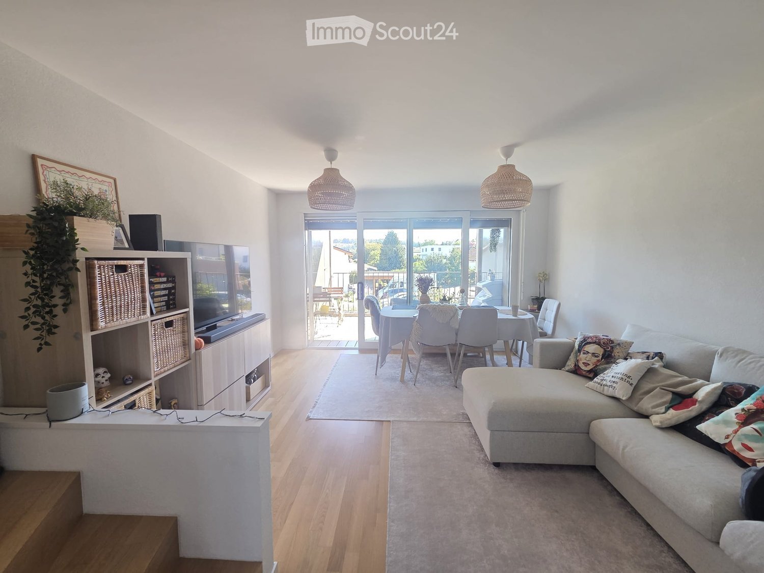 Modern living room with a grey sectional sofa, coffee table, dining area with chairs and table, shelves with decorative items, and large windows leading to a balcony.