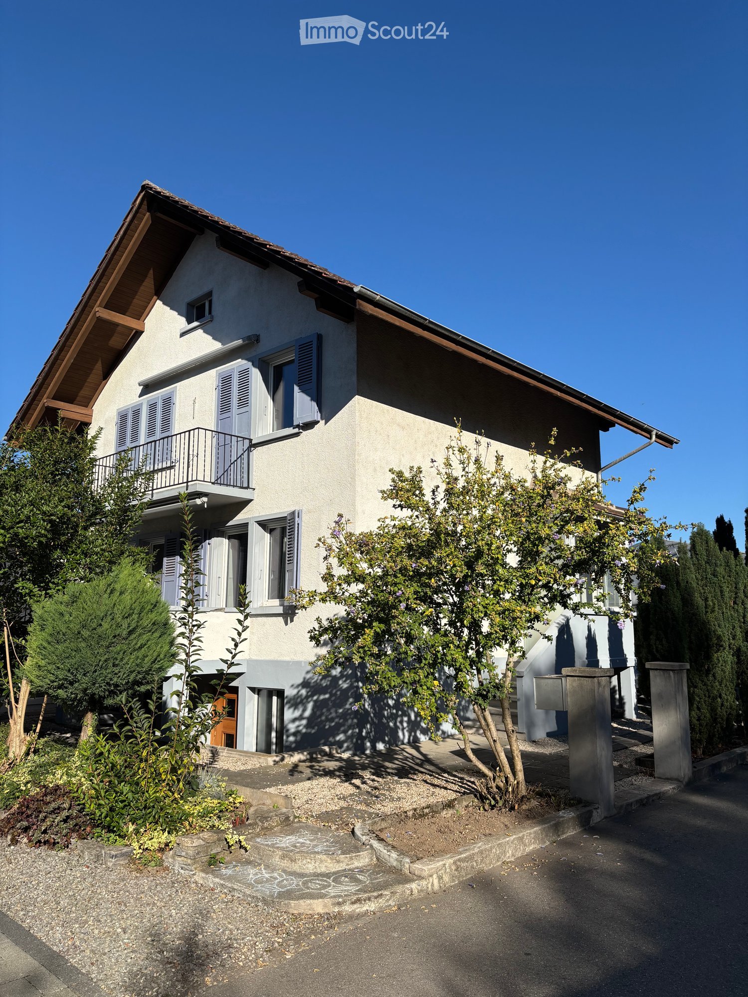 Two-story house with light-colored walls, multiple windows with shutters, a small balcony on the upper floor, and surrounding greenery.
