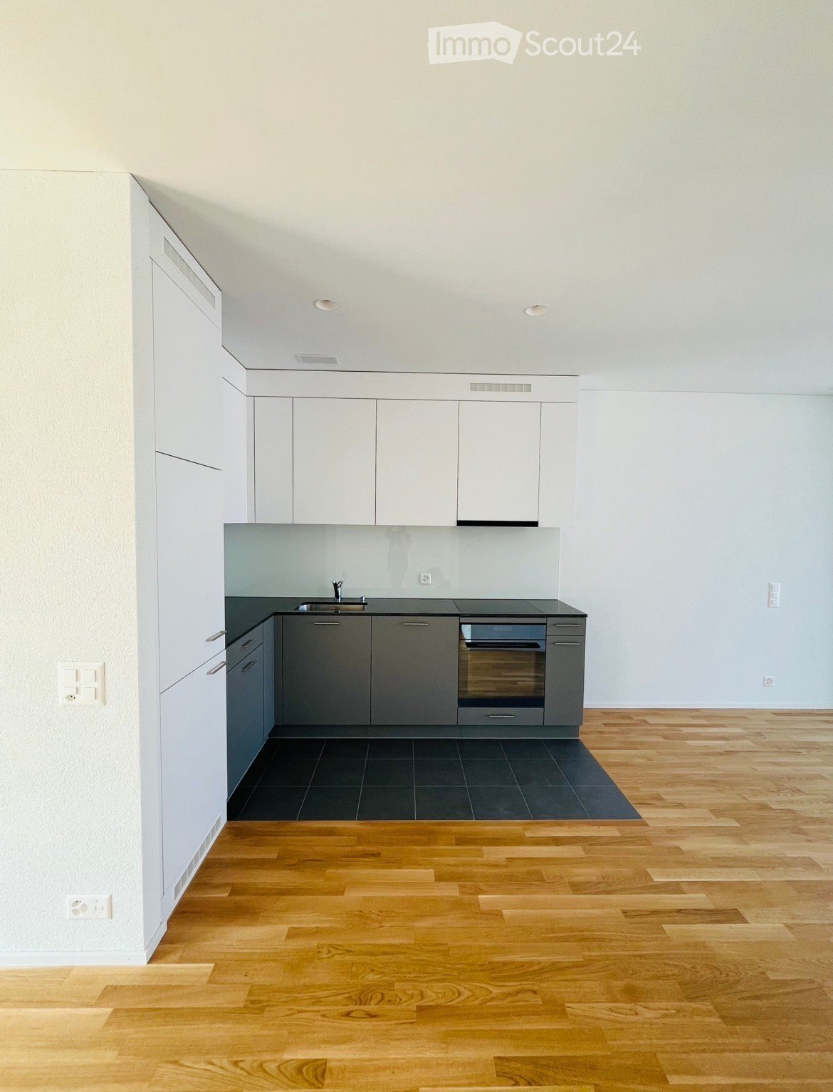 Modern, minimalist kitchen with white and grey cabinetry, a dark countertop, and a wooden floor. The space is empty, with no appliances or furniture visible.