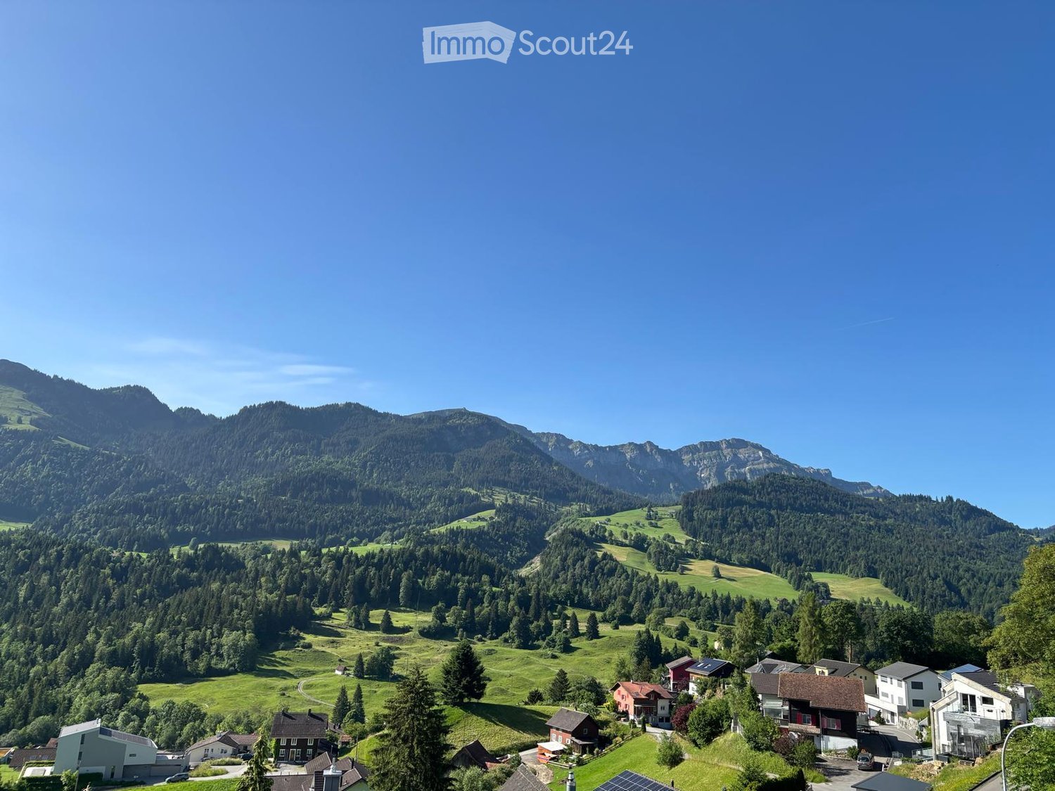 A scenic view of a village nestled in a lush green valley surrounded by mountains. The village features several houses with varied architectural styles, surrounded by dense forests and open fields.