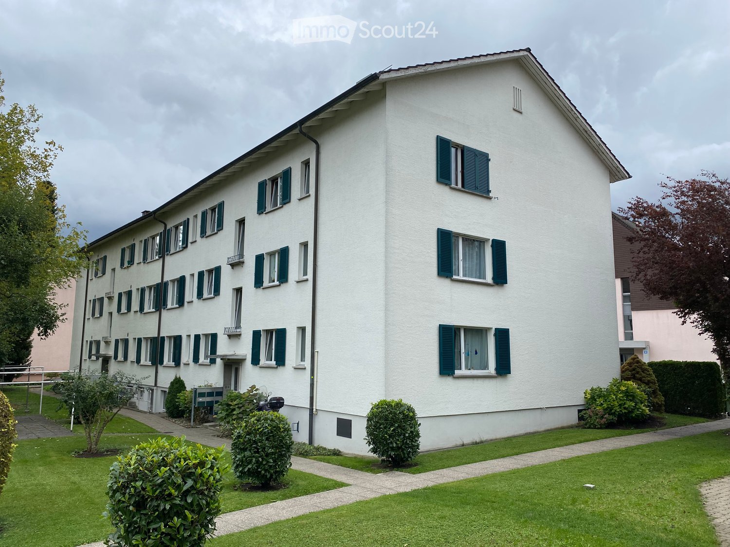 A multi-story white building with green shutters and multiple windows, surrounded by a well-maintained lawn and small shrubs.