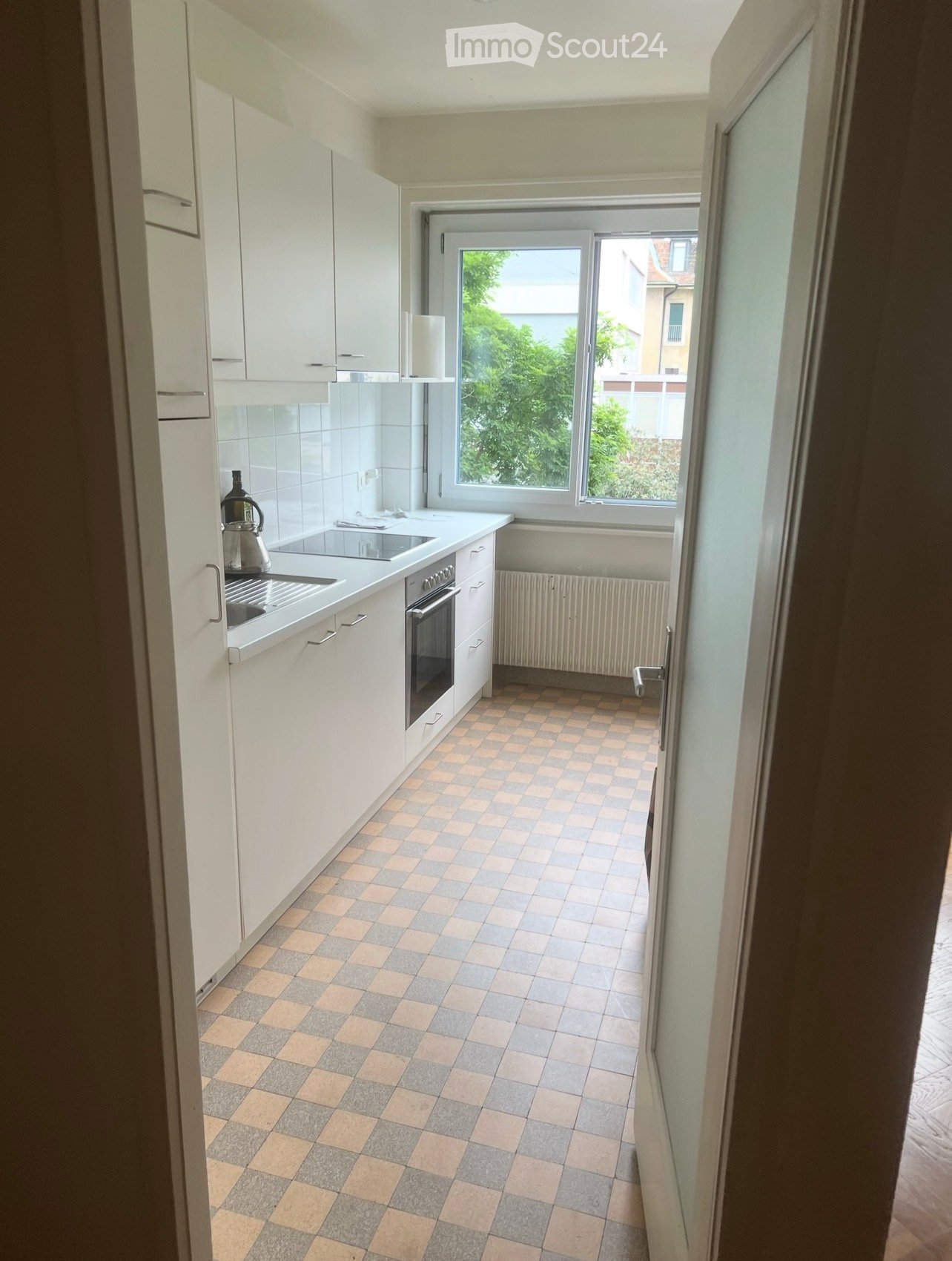 Modern kitchen with white cabinets, a tiled floor, and a window overlooking greenery. Includes a sink, oven, and teapot on the counter.