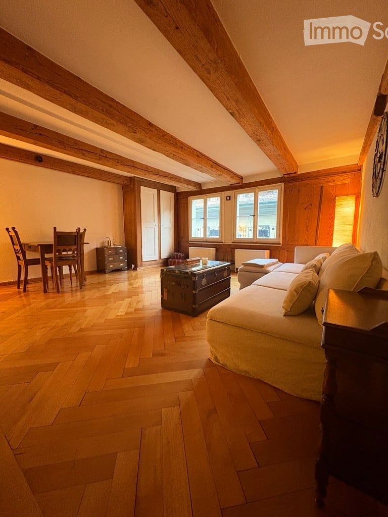 Cozy attic living room with wooden beams, hardwood floor, large windows, sofa, coffee table, dining area with table and chairs, and a small cabinet.