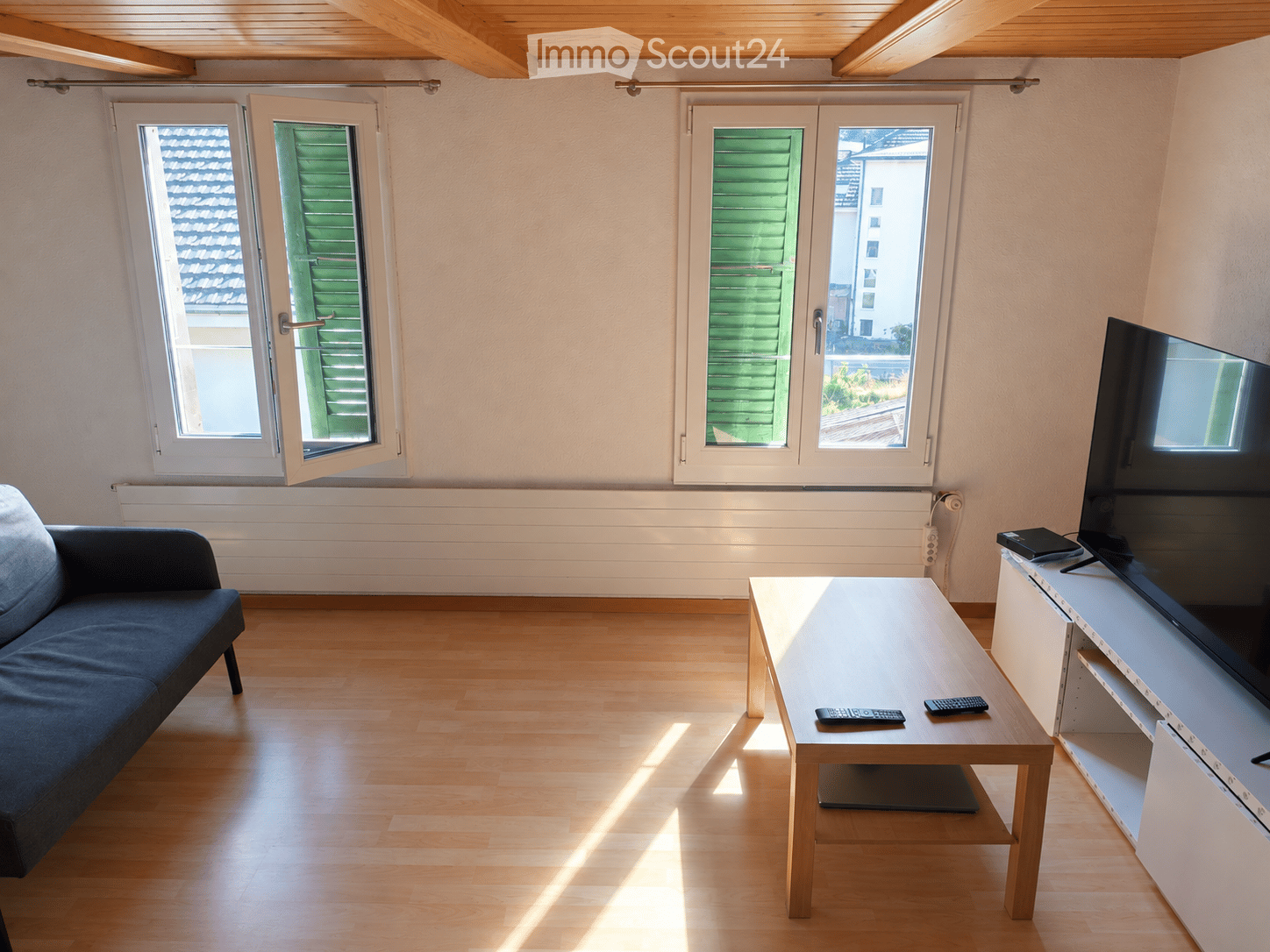 Bright living room with wooden flooring, large windows, a dark blue sofa, a light wooden coffee table with remote controls, and a TV on a white stand.