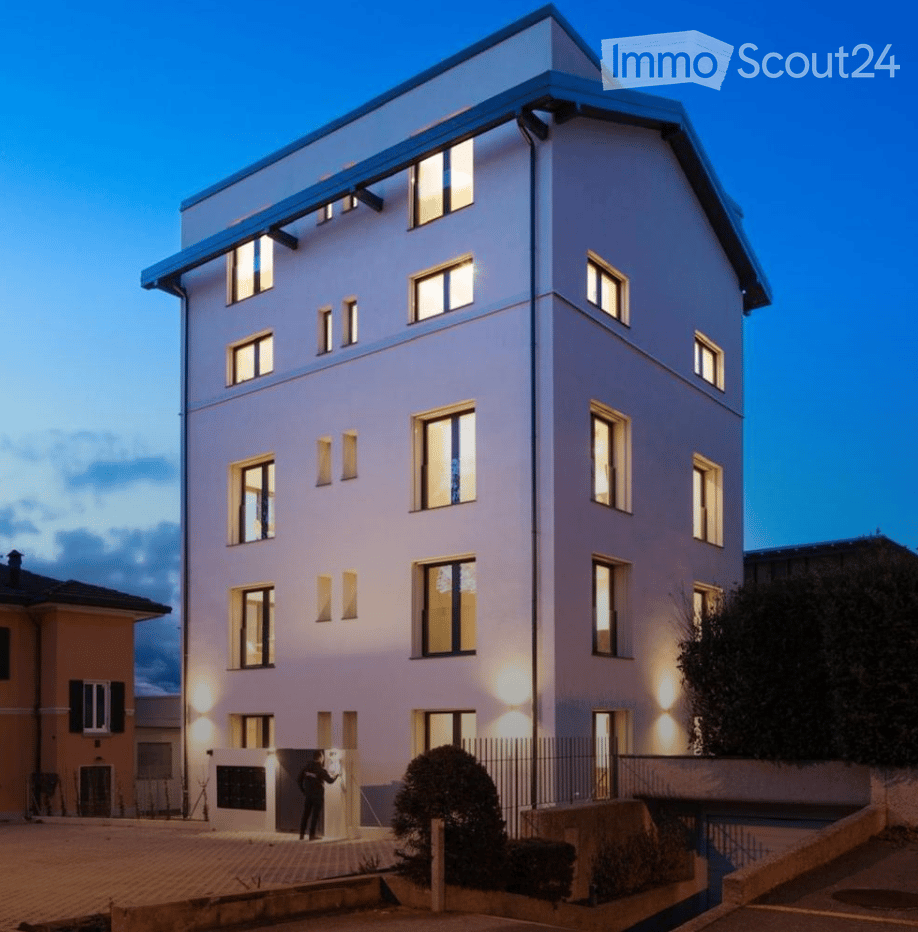 Modern multi-story residential building with illuminated windows and clean lines, featuring a raised ground floor and contemporary design.