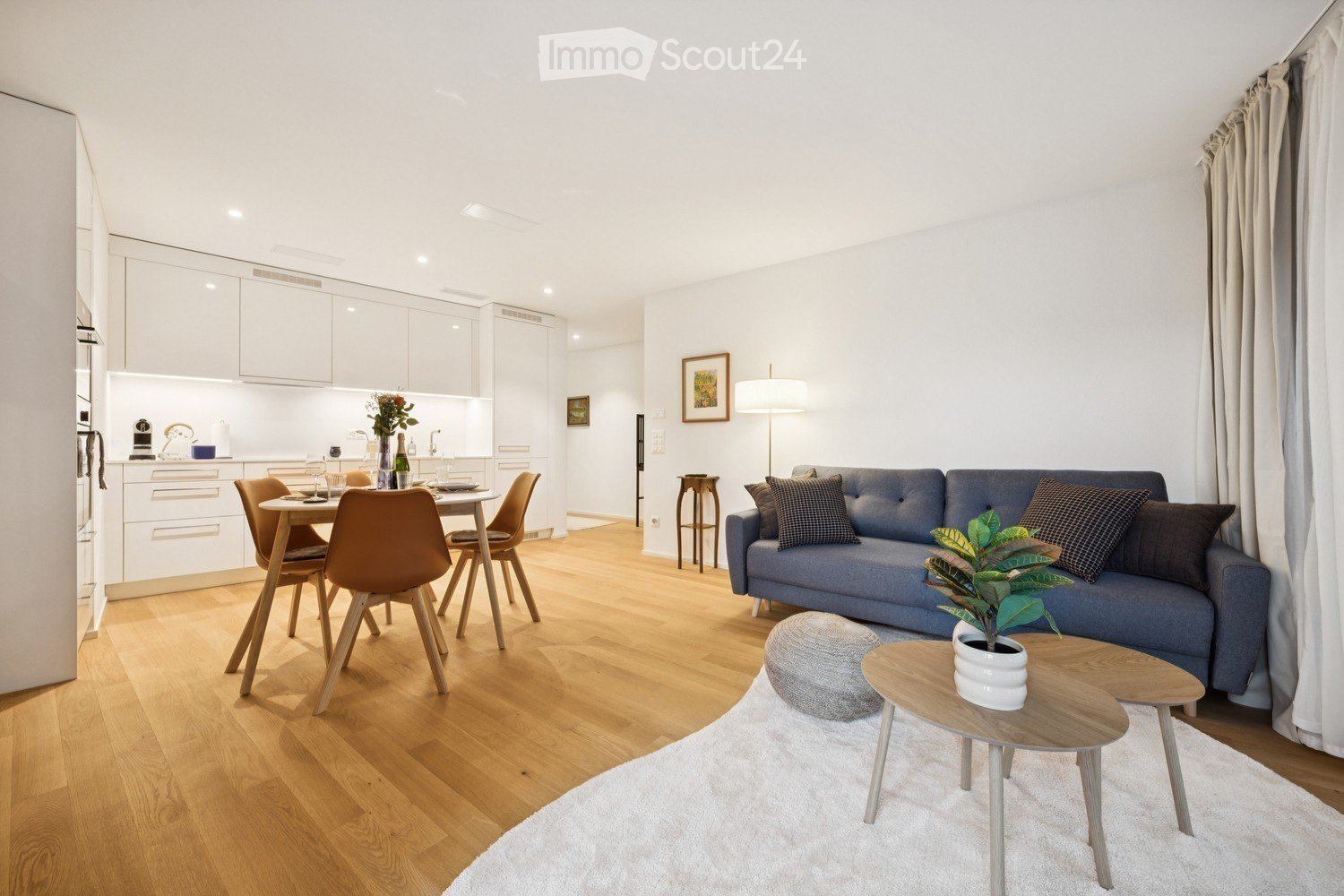 Modern open-plan living and dining area with light wood flooring, white walls, and large windows. Features a blue sofa with patterned cushions, a small coffee table with a potted plant, a grey ottoman, a dining table with four chairs, and a fitted kitchen