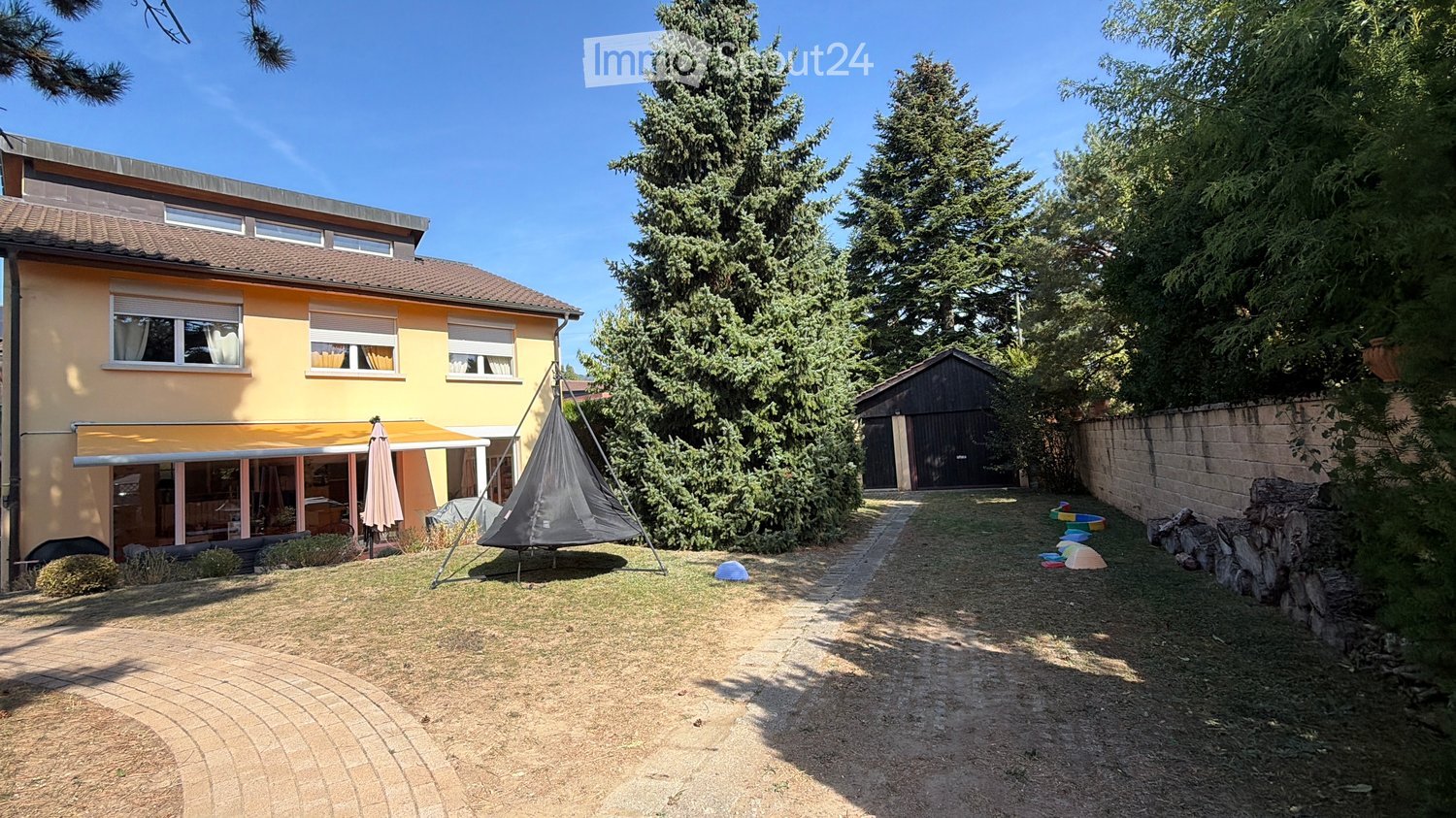 A sunny garden area with a hammock, a covered patio, and a small shed. The house is yellow with multiple windows and a garage.