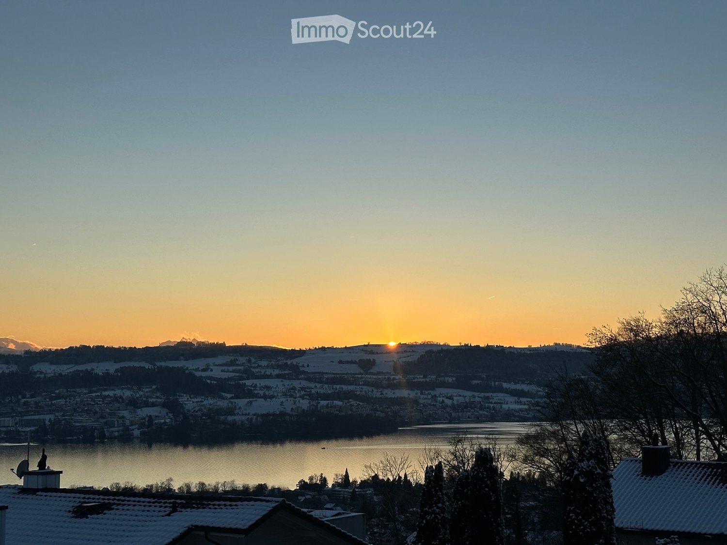 Panoramic view of a snow-covered landscape at sunset, featuring a lake, distant mountains, and rooftops in the foreground.