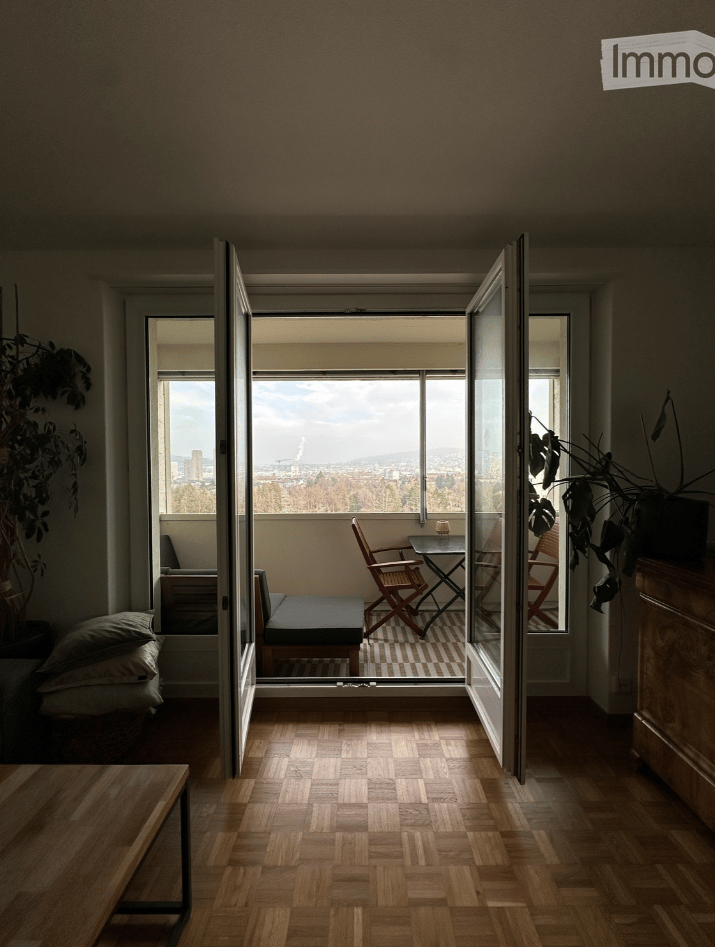 Modern living room with wooden flooring, large windows offering a city view, and a small seating area with a sofa, table, and chairs.