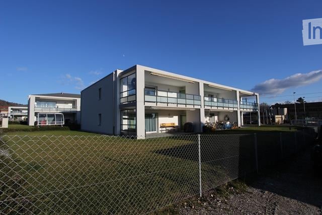 Modern two-story building with white exterior, multiple windows, and balconies. The building is surrounded by a fenced area with grass and gravel.