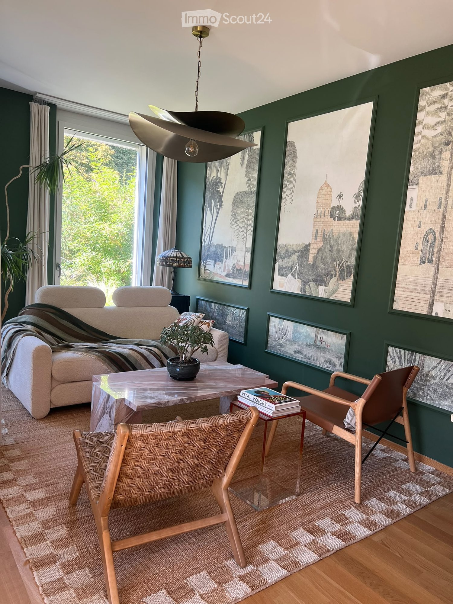Living room with green walls, large windows, a beige sofa, a glass coffee table, two chairs, a table lamp, and artwork on the walls.