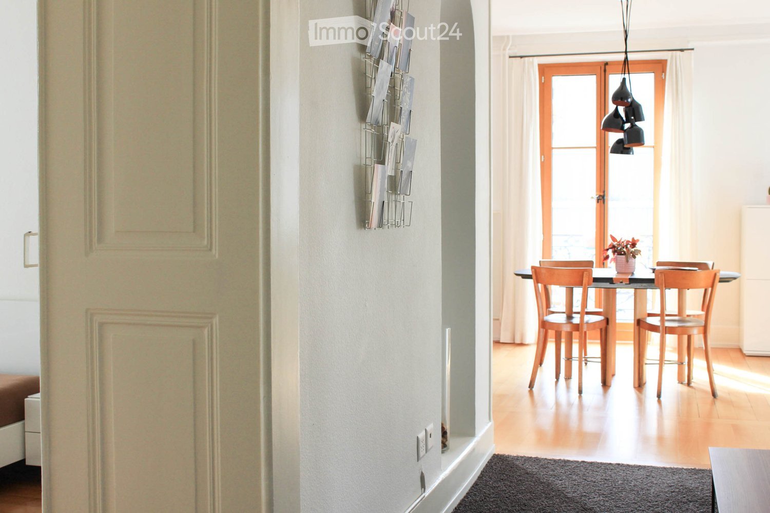 A well-lit hallway with light-colored walls, a wooden floor, and a small area leading to a dining space with a table and chairs.