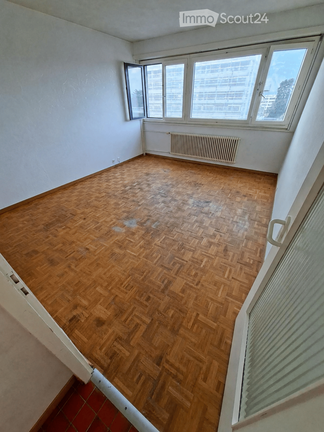 Empty room with wooden herringbone flooring, white walls, large windows, and a radiator. No furniture or equipment present.